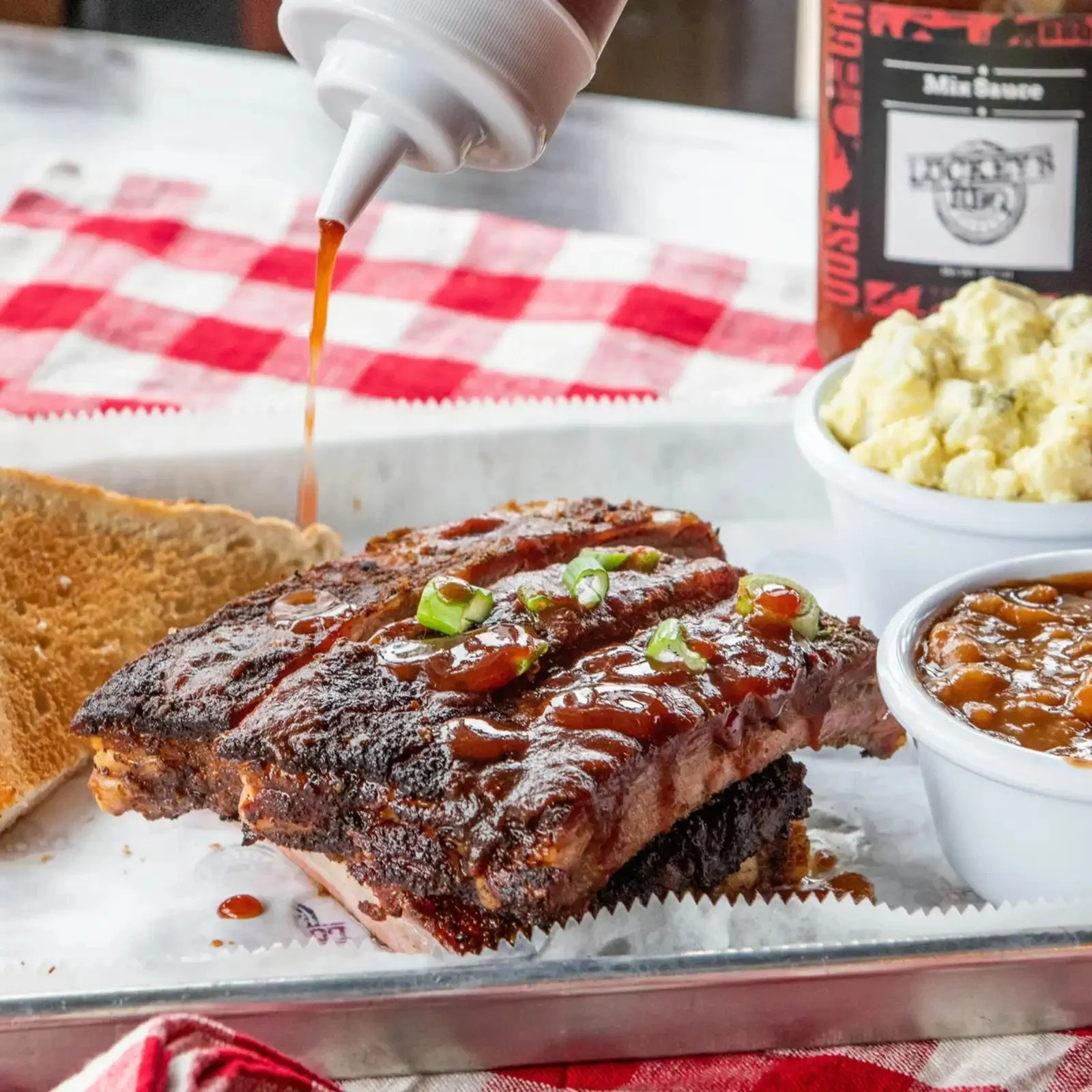 Barbecue ribs with green onions, served with side dishes of mashed potatoes, baked beans, and toast, with a bottle of barbecue sauce in the background.