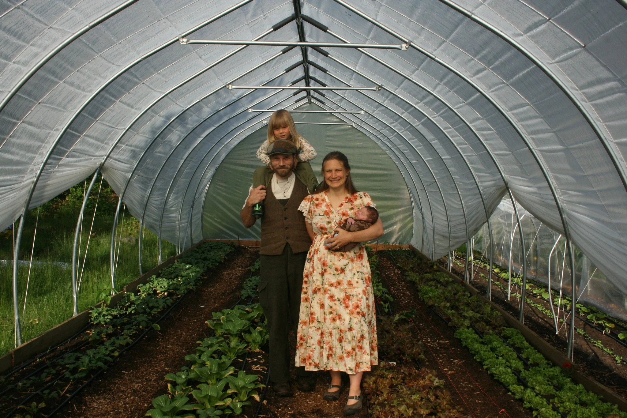 Family of four, including a man, woman, young boy, and newborn, standing inside a greenhouse with rows of leafy vegetables.