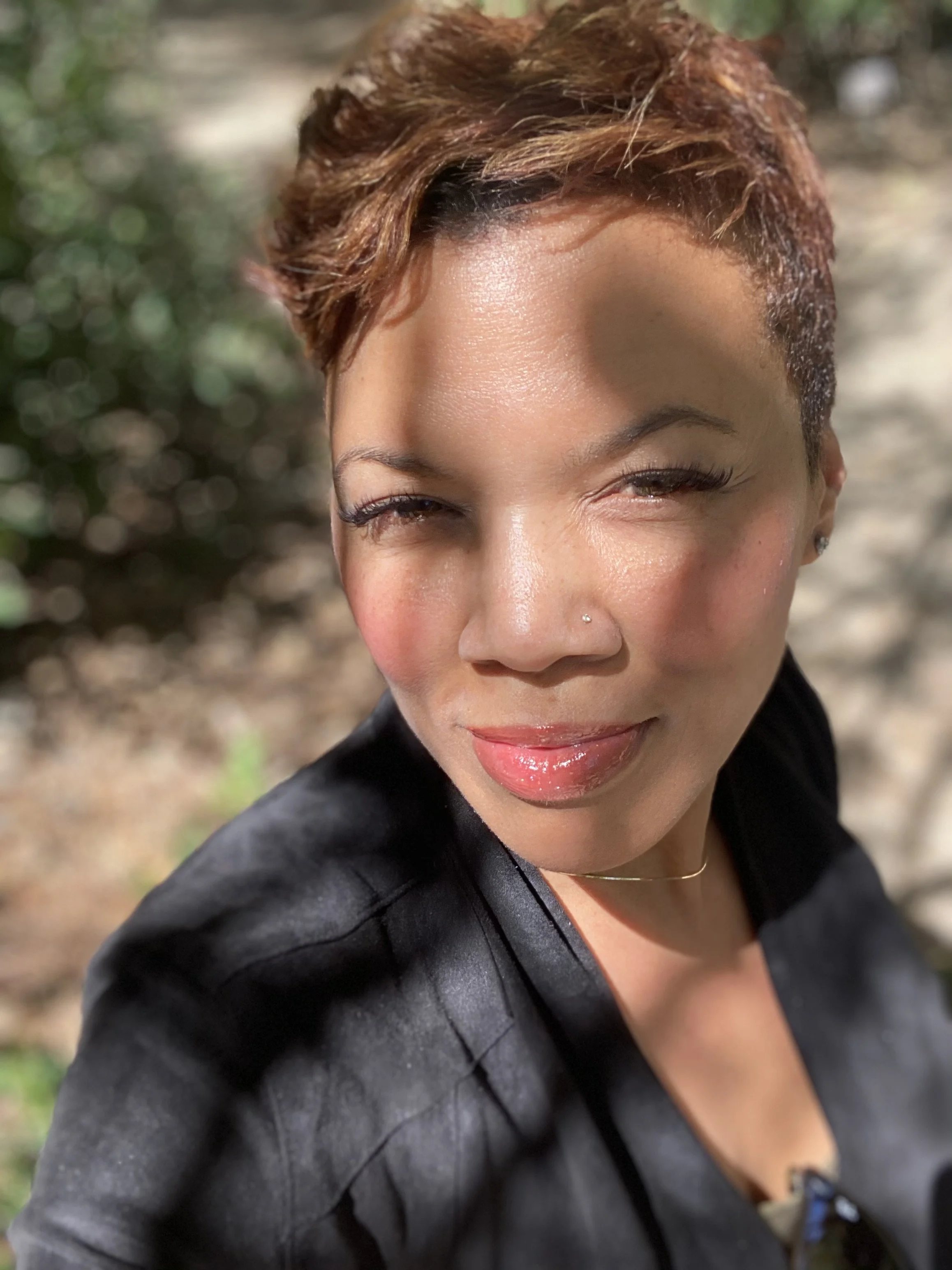 A woman with short, wavy, reddish-brown hair, wearing a black leather jacket, smiling with makeup, outdoors with blurred trees in the background.