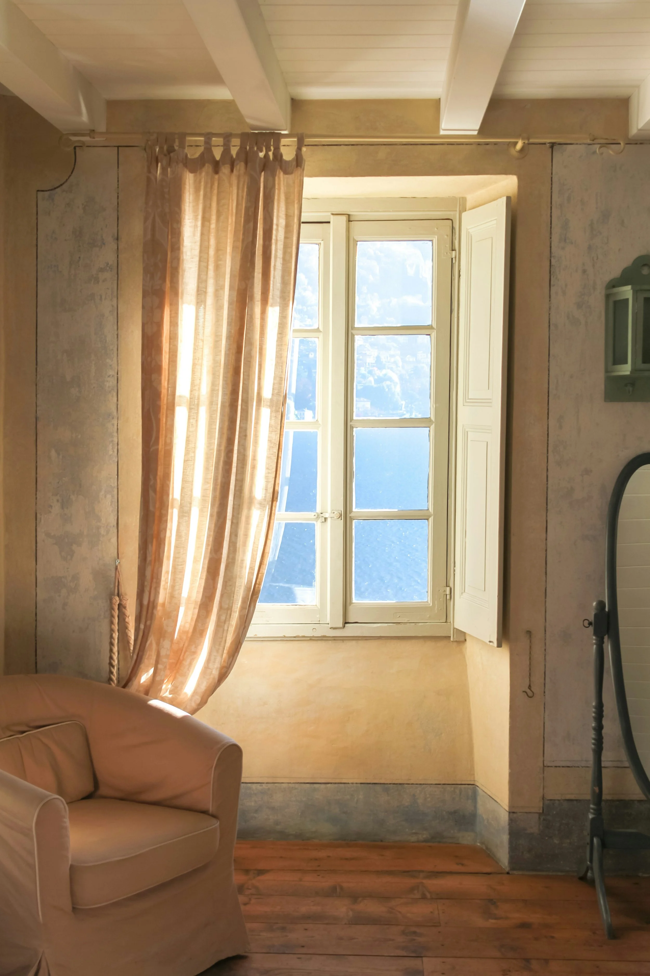 A cozy room with a wooden floor and a beige armchair in the corner. There is a window with open shutters and a sheer, patterned curtain letting in sunlight. The room has a rustic, vintage feel with textured walls.