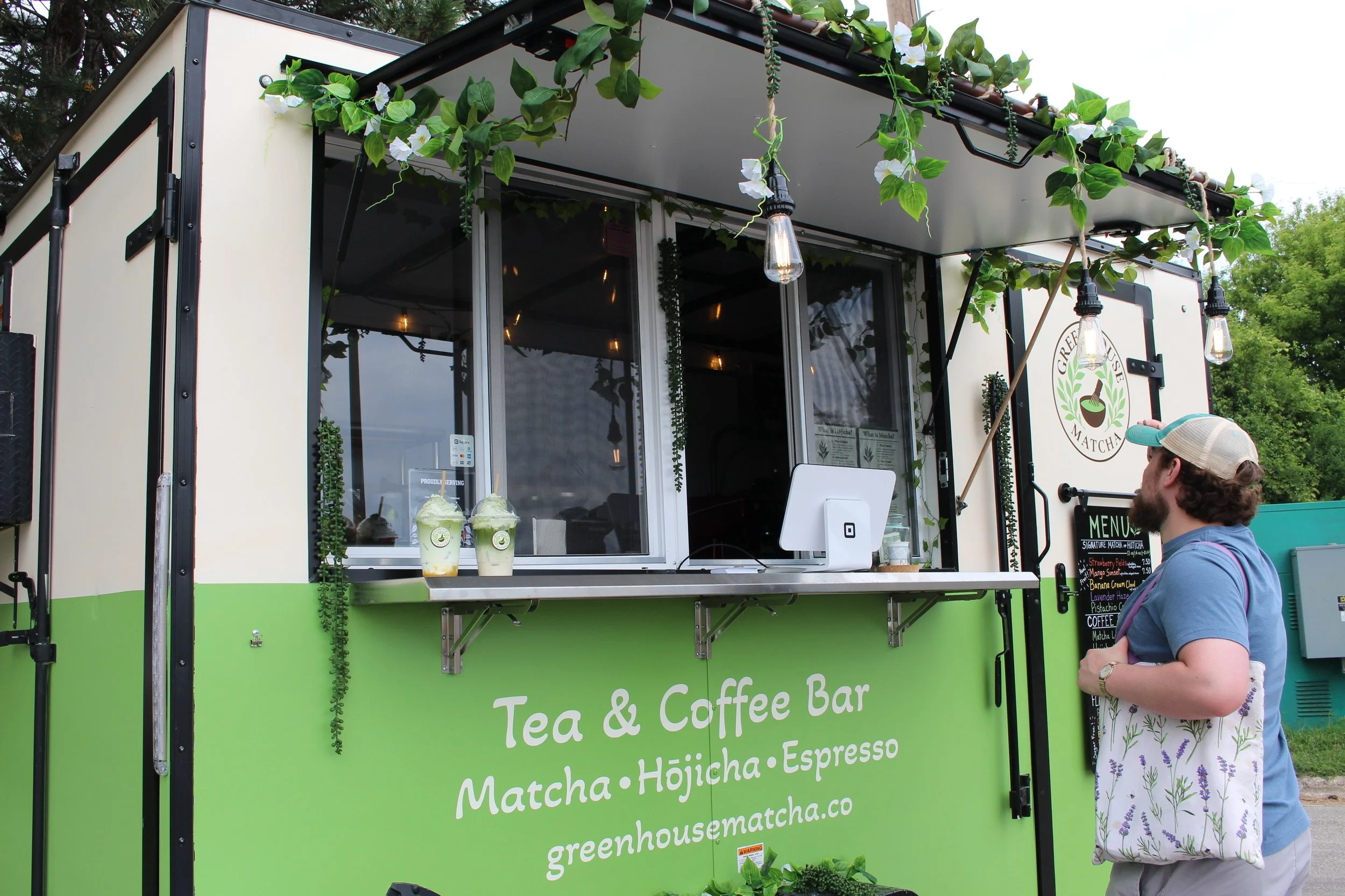 Green food truck with sign advertising tea and coffee bar, serving matcha, hojicha, and espresso, with two cups of green tea or matcha drinks on the counter, and a person in a hat and floral skirt standing nearby.