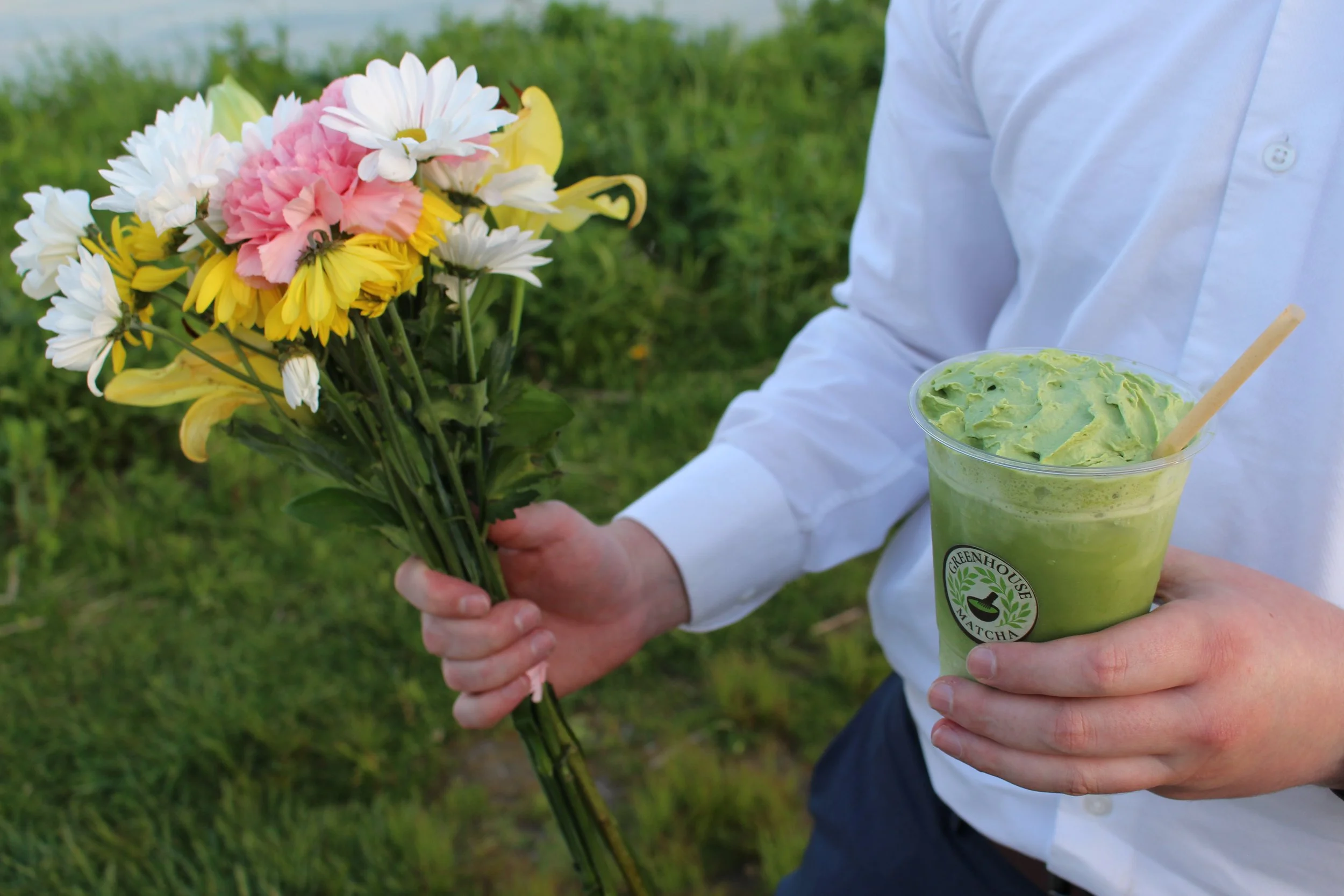 Person in white shirt holding a bouquet of pink, white, yellow, and peach flowers in one hand and a green matcha beverage in a clear cup in the other hand, outdoors on grass.