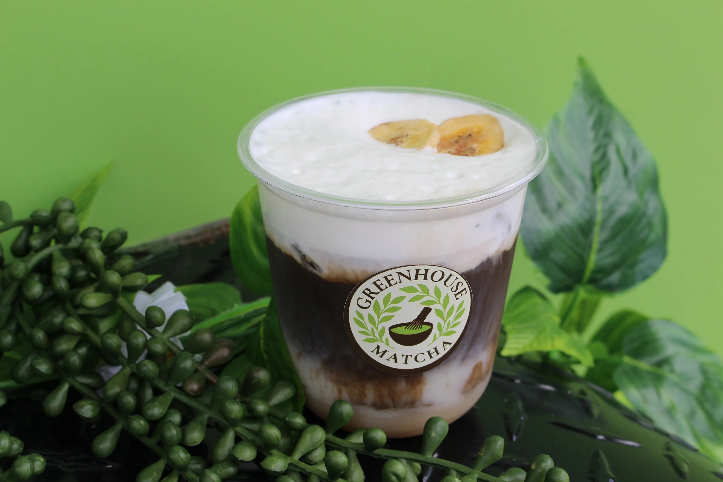 A matcha drink with whipped cream and banana slices in a clear plastic cup, labeled 'Greenhouse Matcha', surrounded by green leaves and a cluster of small green berries against a green background.