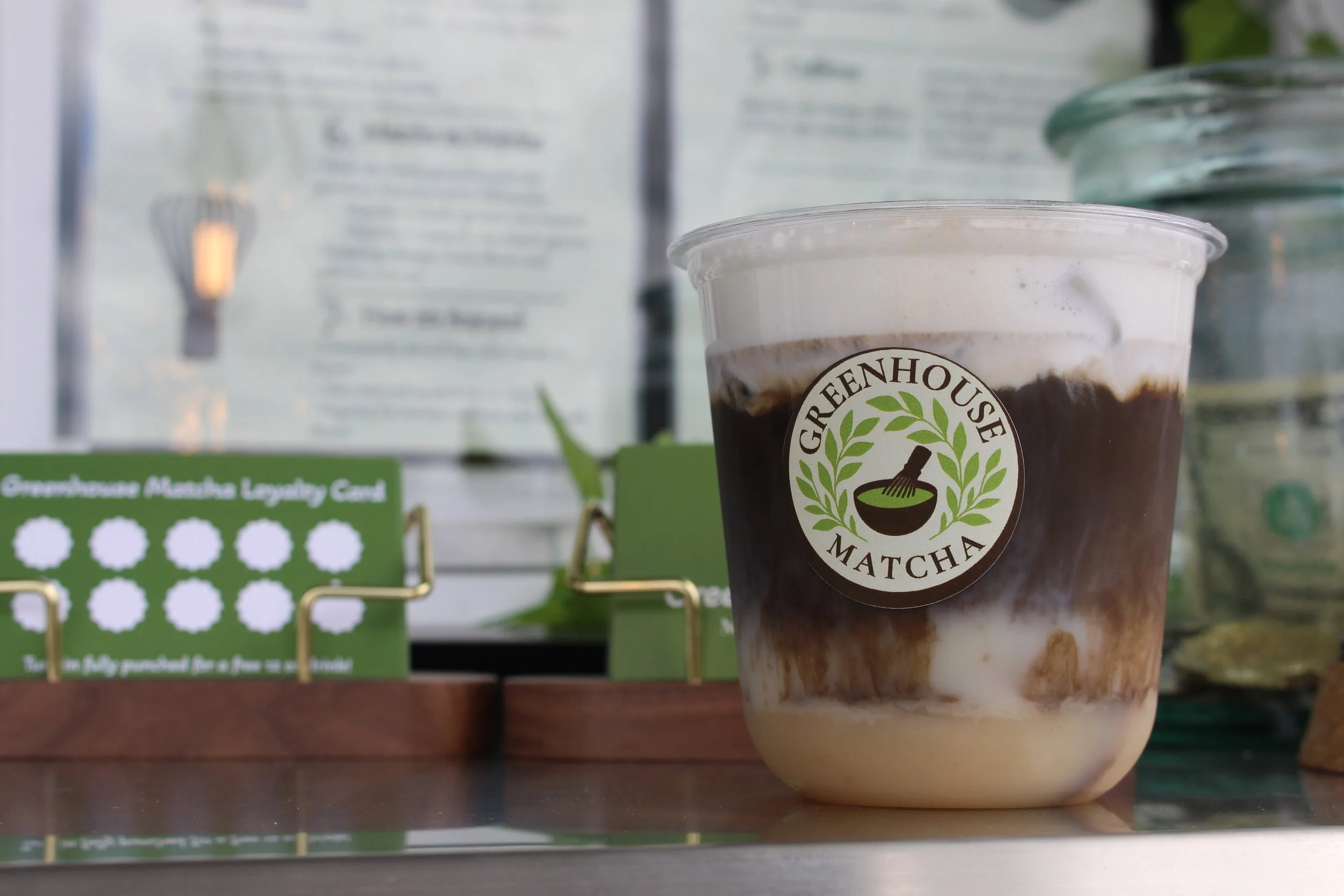 A plastic cup of iced matcha latte with whipped cream, sitting on a wooden surface, with a logo that reads 'Greenhouse Matcha' featuring a green leaf and bowl of matcha.