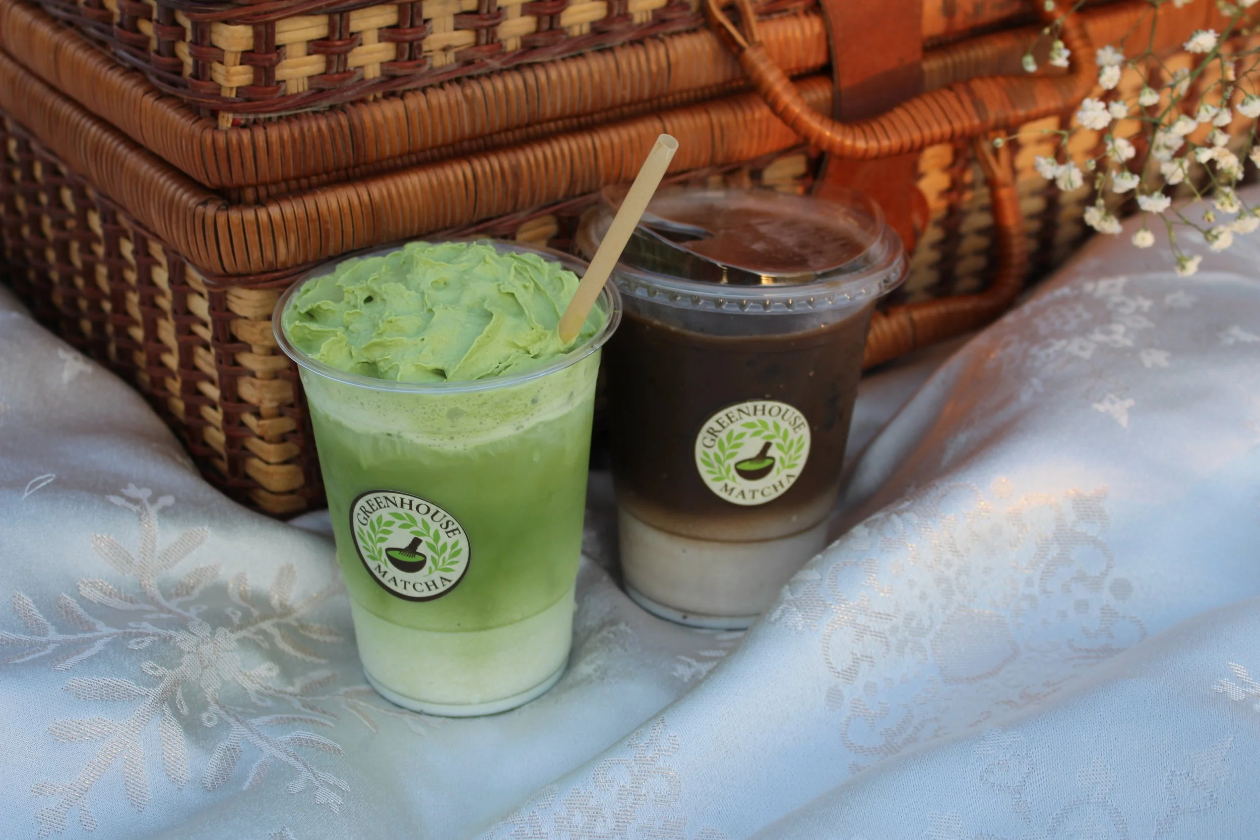 Two cups of beverages on a picnic blanket with a wicker basket in the background. One drink is green, topped with a thick layer of matcha-flavored foam. The other is dark brown hojicha with a clear plastic lid and straw, labeled "Greenhouse Matcha."