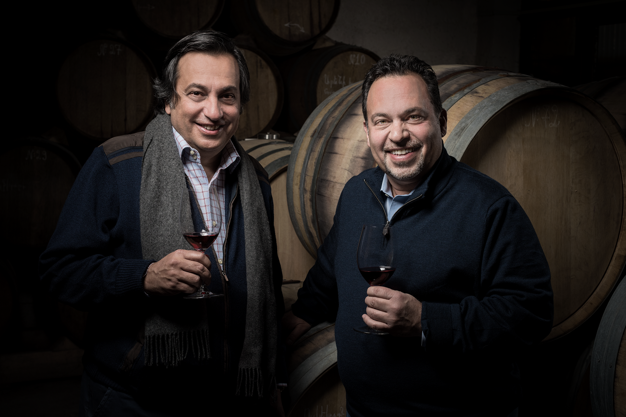Brothers Vahe and Vigen Yacoubian holding glasses of red wine in a cellar with wine barrels in the background.