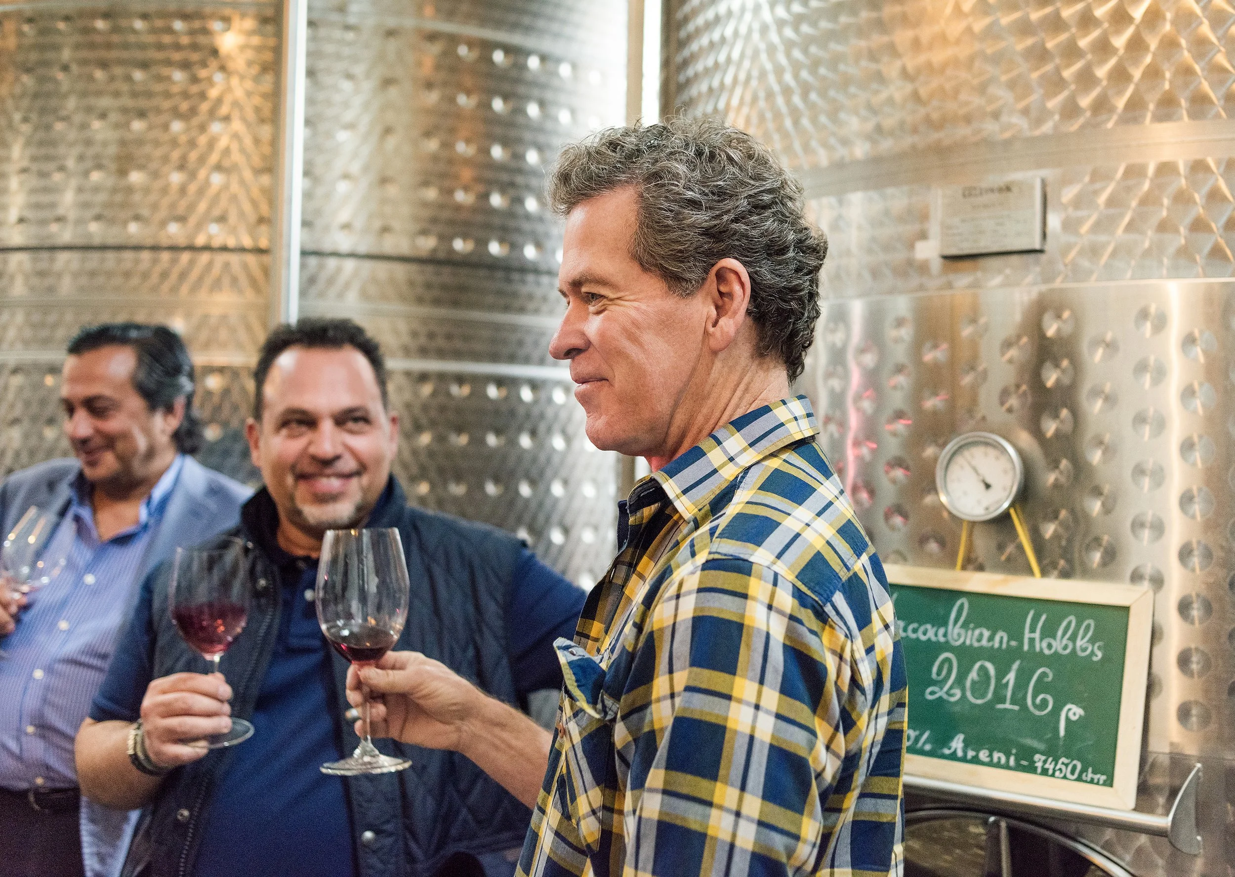 Vahe and Vigen Yacoubian with Paul Hobbs at a winery in front of large stainless steel fermentation tanks, holding glasses of red wine, smiling and socializing.