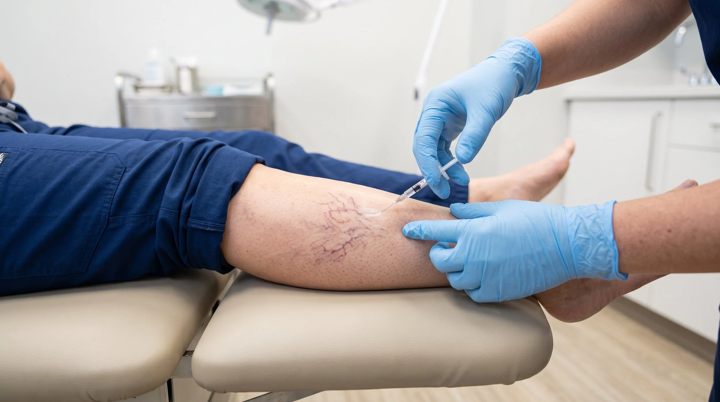 A healthcare professional in blue gloves administering an injection into a patient's leg, which has visible varicose veins, in a medical setting.