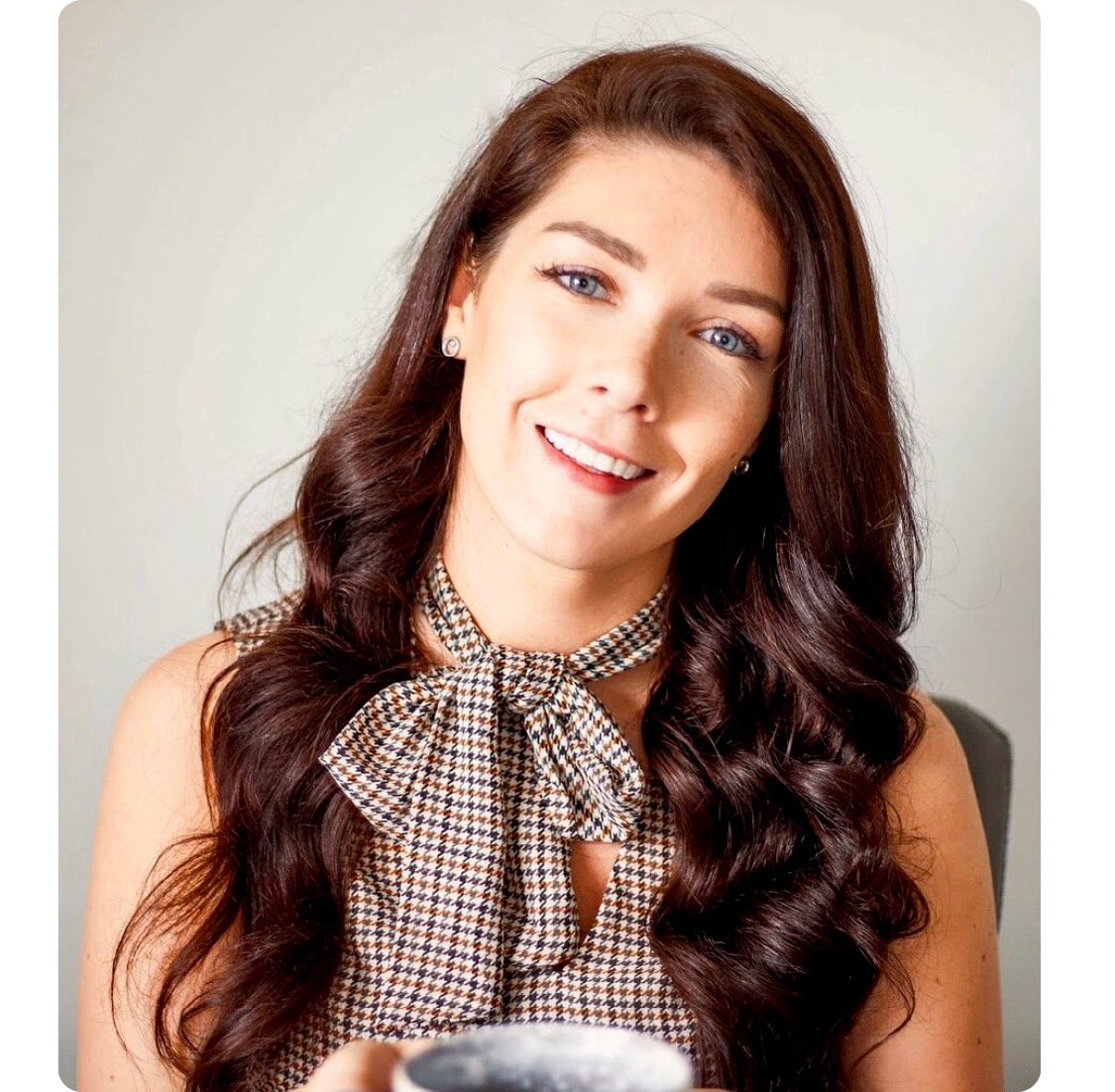 A young woman with long, wavy brown hair smiling and looking at the camera, wearing a checkered sleeveless top with a bow tie around her neck, earrings, and holding a cup.
