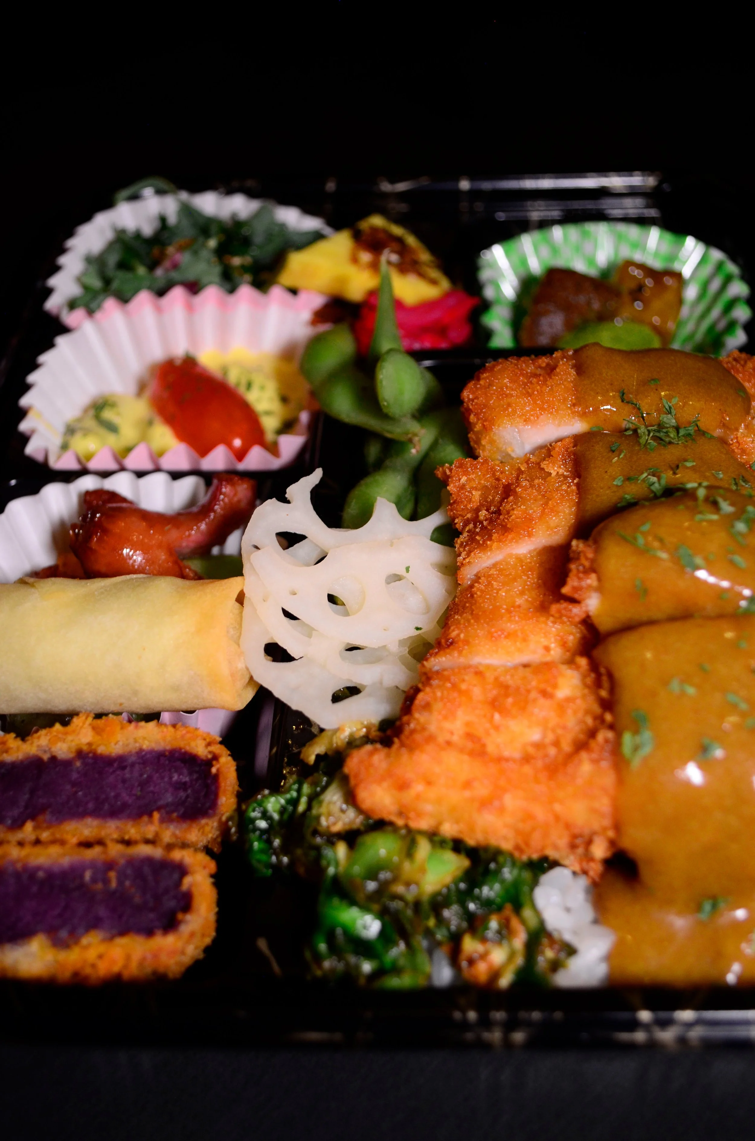 A close-up of a Japanese bento box with fried chicken topped with curry sauce, pickled vegetables, lotus root, green beans, purple sweet potato croquettes, and assorted side dishes in colorful paper cups.