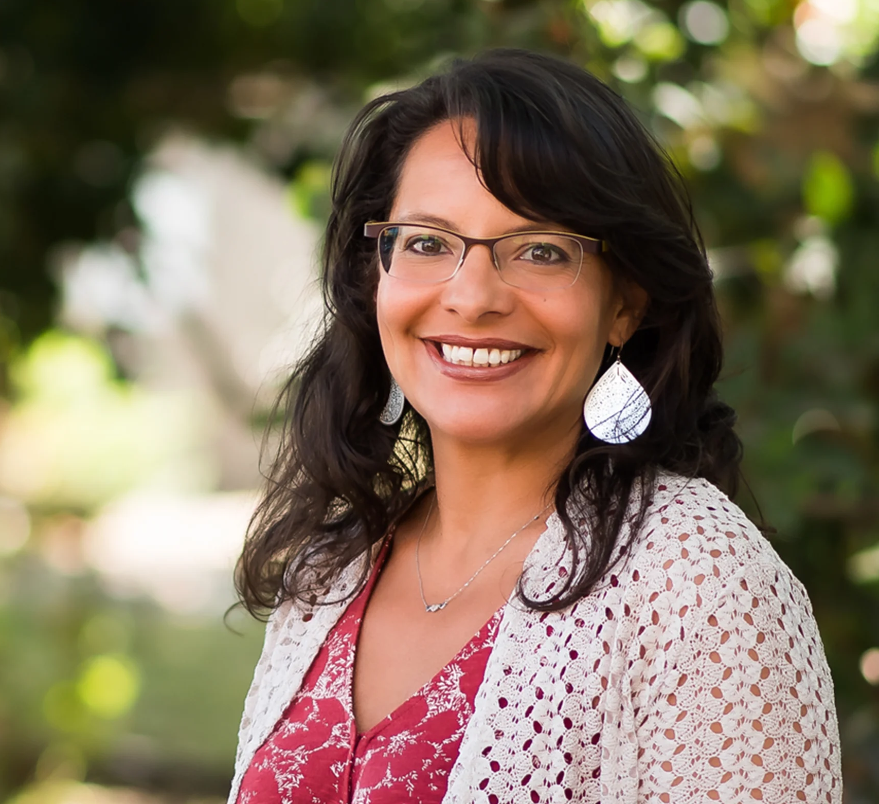 A smiling woman with dark hair and glasses, standing outdoors with green foliage in the background.