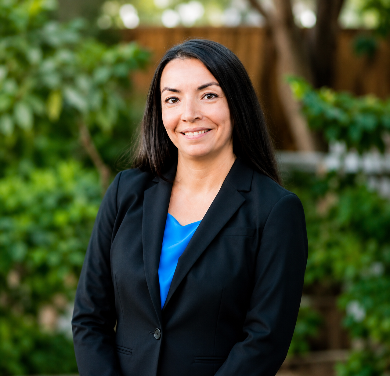 A woman with long dark hair, wearing a black blazer over a blue top, smiling outdoors with greenery in the background.