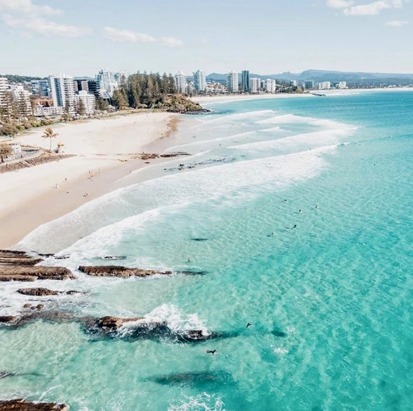 A coastal city coolangatta beach with sandy shores, turquoise water, waves, beachgoers, and modern high-rise buildings in the background under a partly cloudy sky.