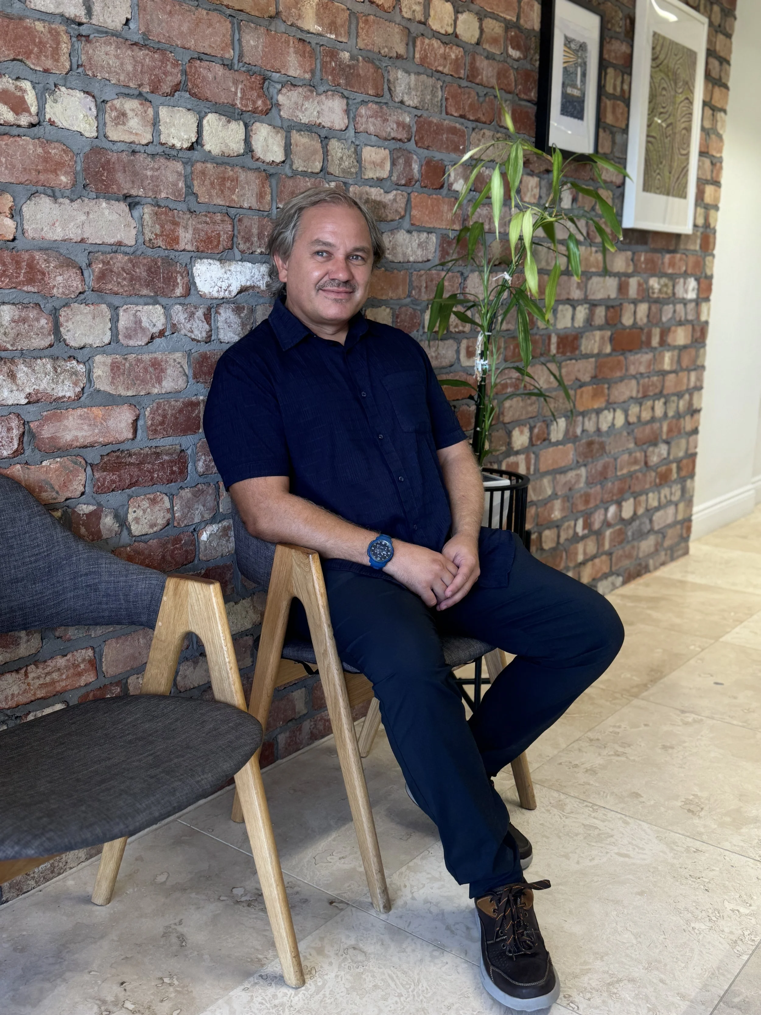A man sitting on a wooden chair against a brick wall, next to a potted plant and framed artwork.