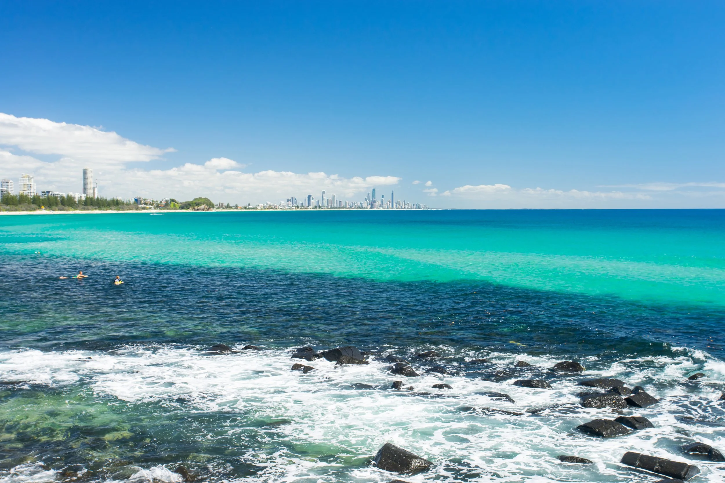 A clear blue sky and turquoise ocean with a city skyline in the distance, rocky shoreline in the foreground, and two people swimming in the water. Burleigh Heads, Gold Coast, Palm Beach