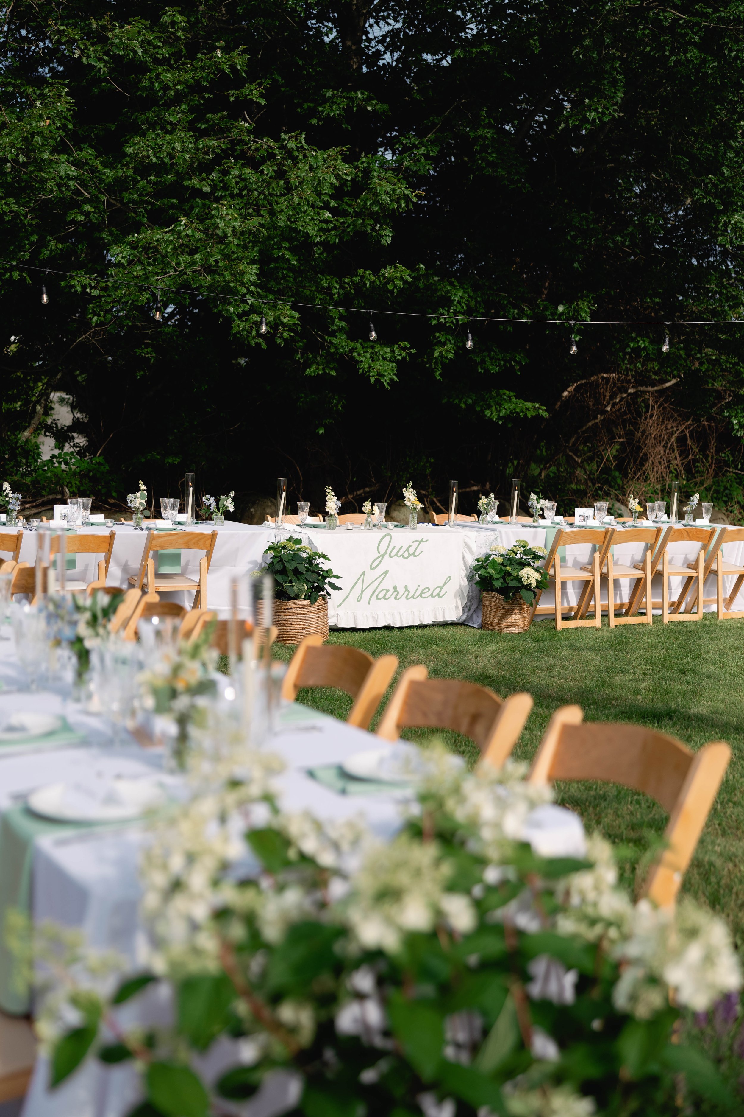 A long outdoor dining table with white tablecloths set with glassware and floral centerpieces, decorated with a