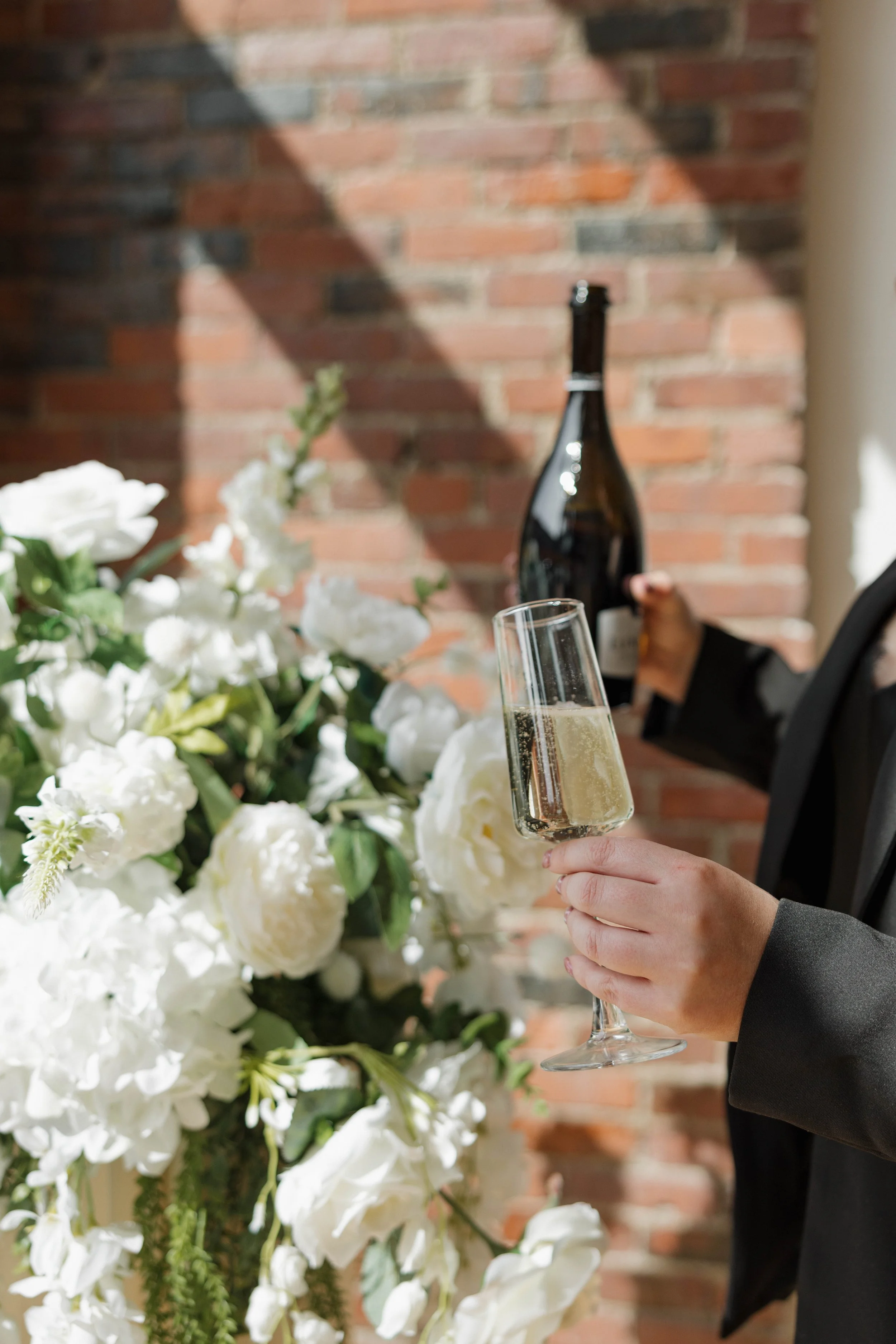 Person in a dark suit holding a glass of champagne, with a floral arrangement of white flowers and green leaves in the foreground, and a person pouring champagne in the background with a brick wall.