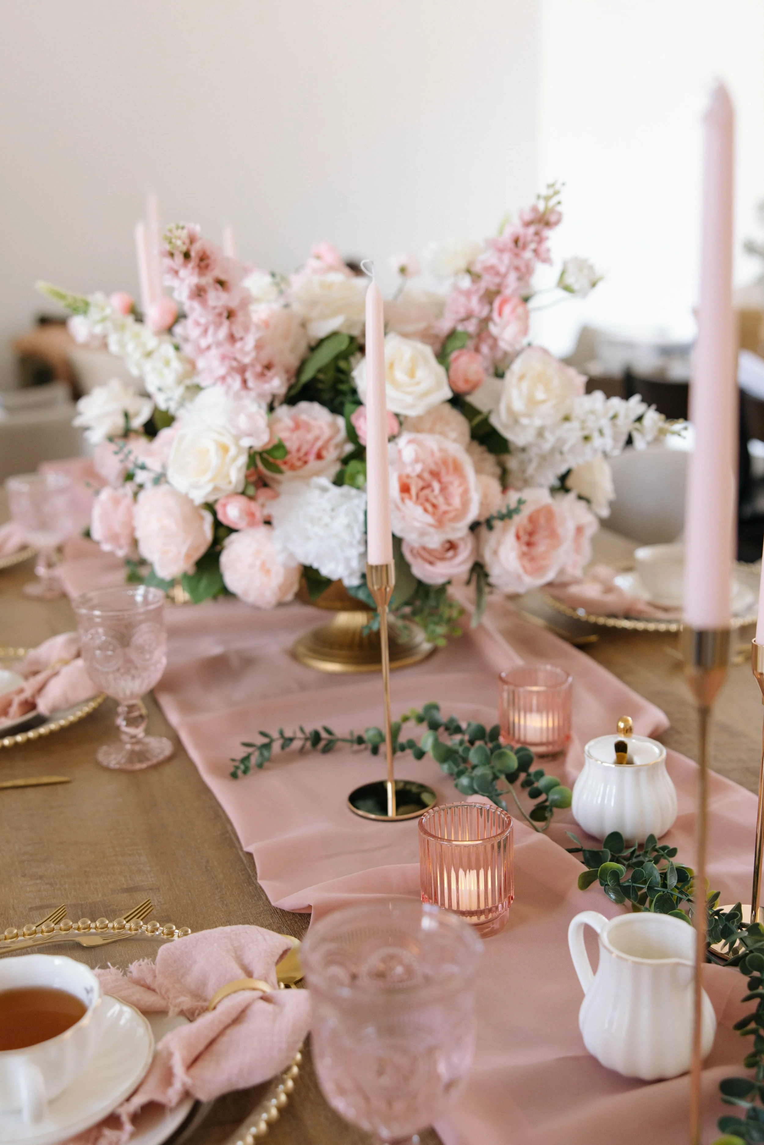 A table decorated with a pink tablecloth, floral centerpiece with white and pink flowers, pink candles, pink glassware, and white teapot, set for a celebration or tea party.