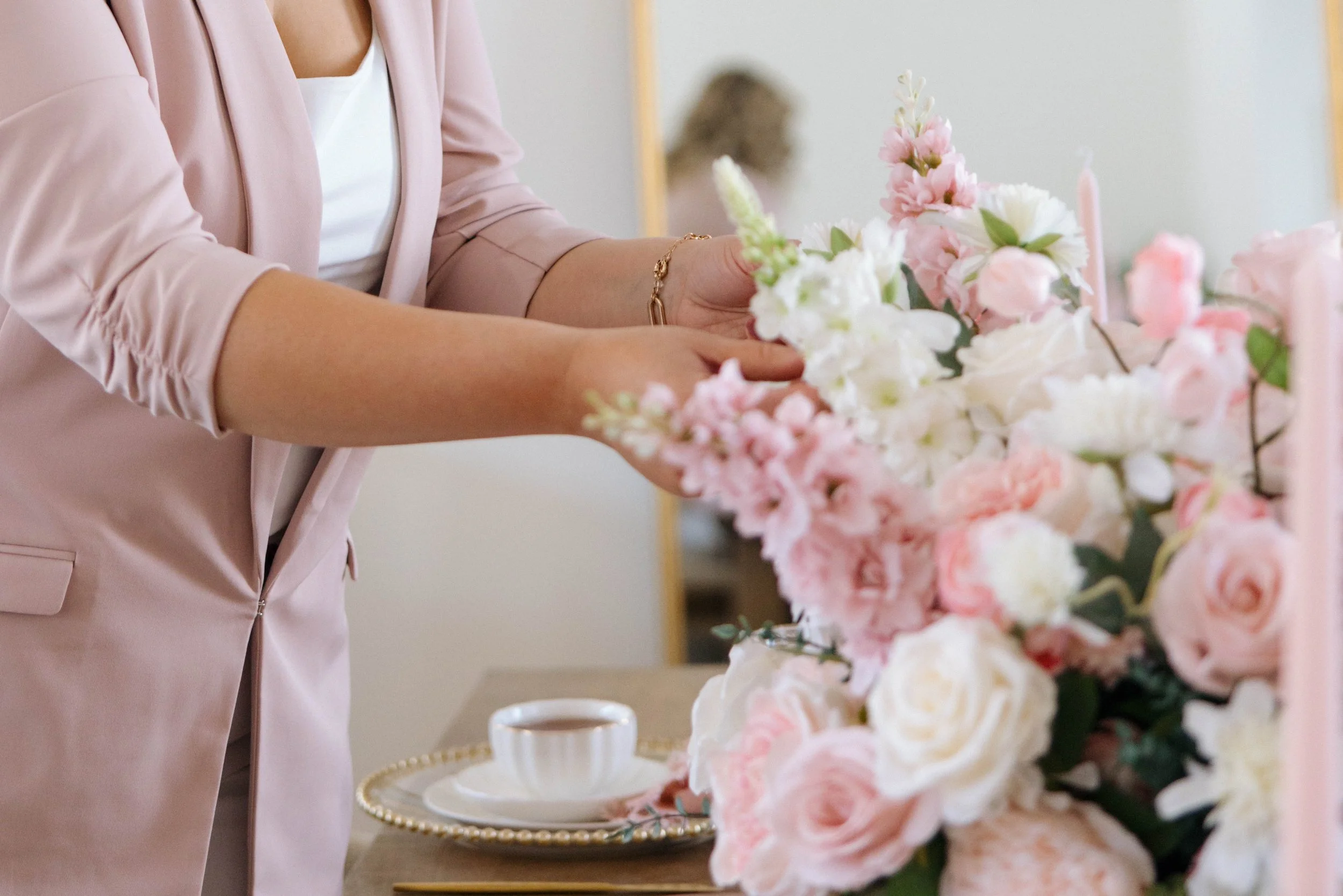 A woman in a pink blazer arranging a bouquet of pink and white flowers on a table with a teacup and saucer.