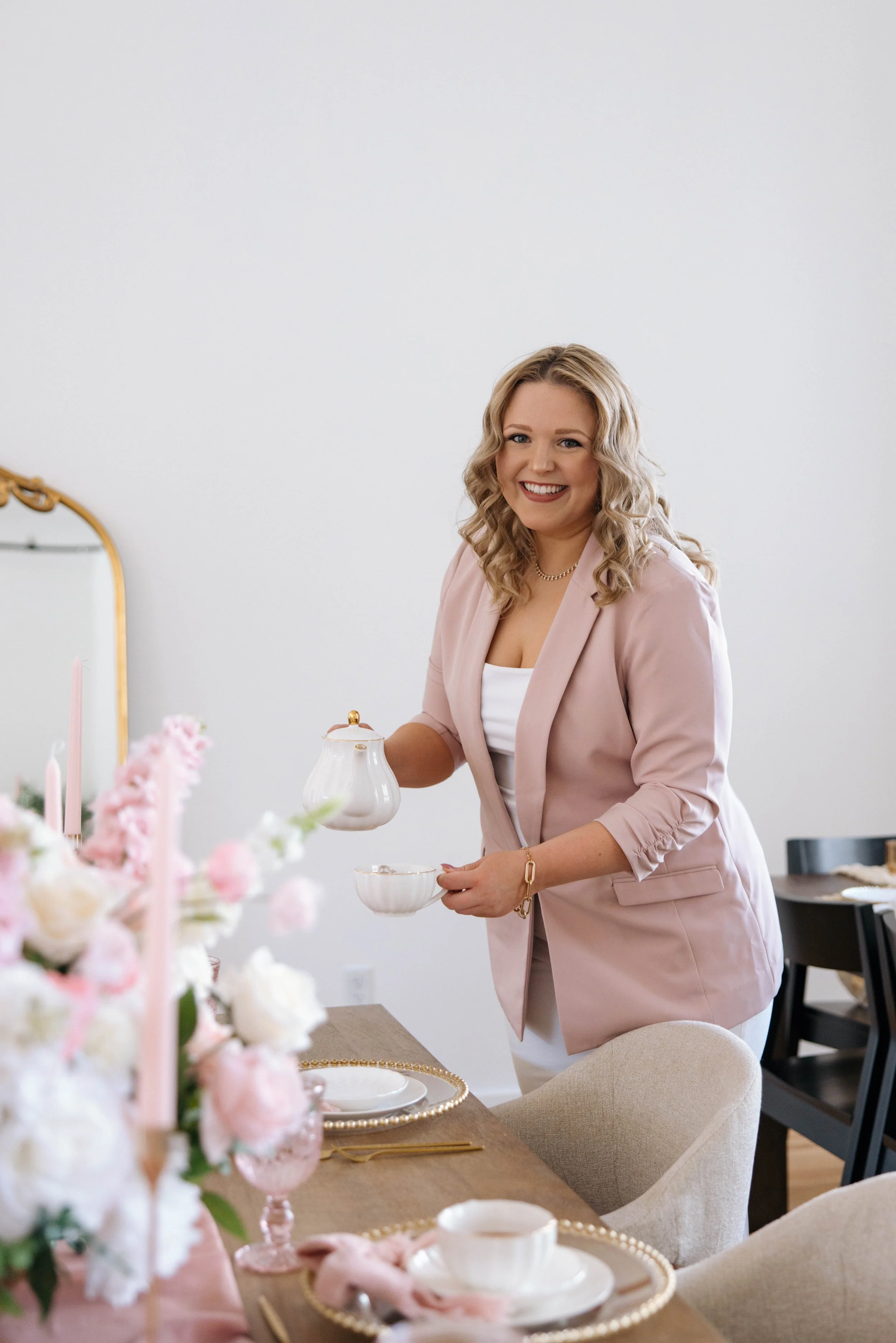 A woman with blonde curly hair, smiling, in a light pink blazer and white outfit, pouring tea at a decorated dining table with pink flowers and gold accents.