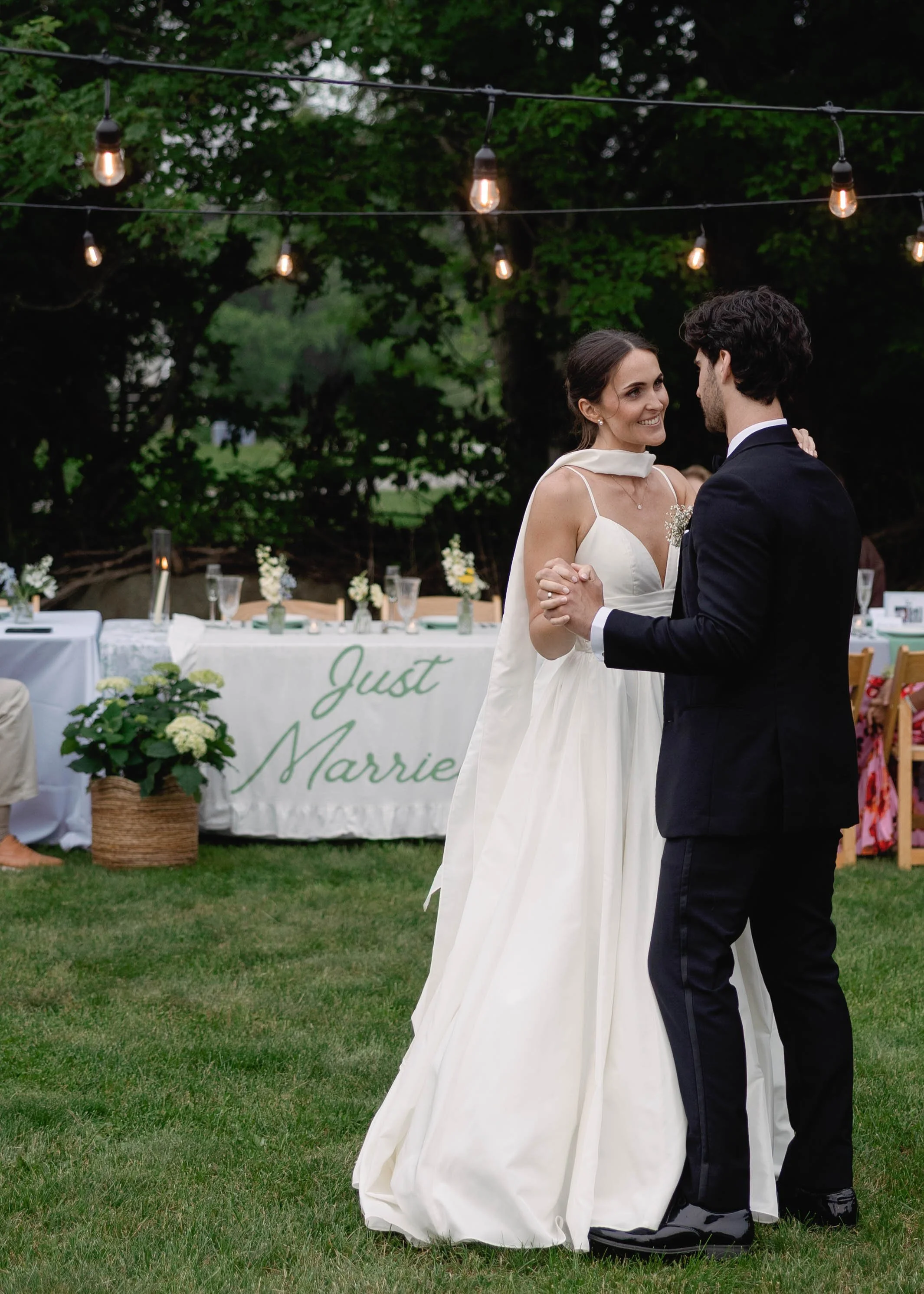 A bride and groom dancing outdoors at their wedding reception under string lights, with a decorated table and greenery in the background.