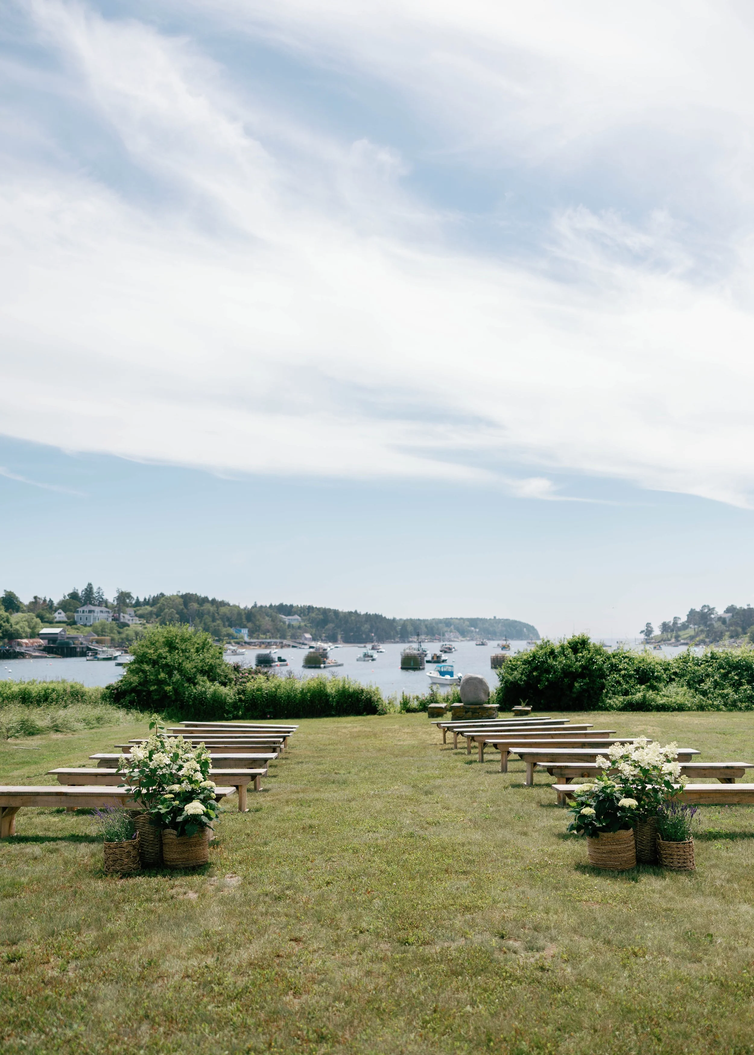 A scenic outdoor setting overlooking a river with boats, white wooden benches, potted flowers, greenery, and a partly cloudy sky.