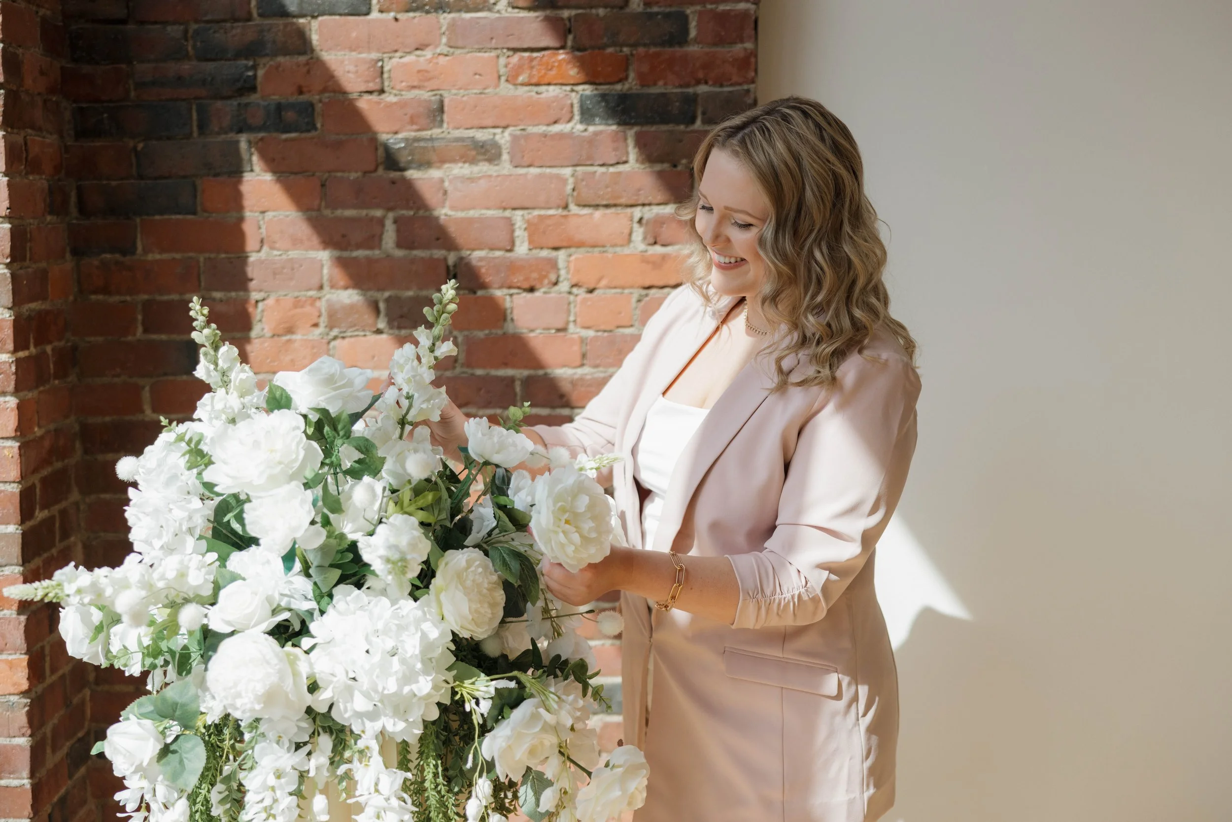 A woman with curly hair, dressed in a light pink blazer and white top, arranging a large bouquet of white flowers near a brick wall.