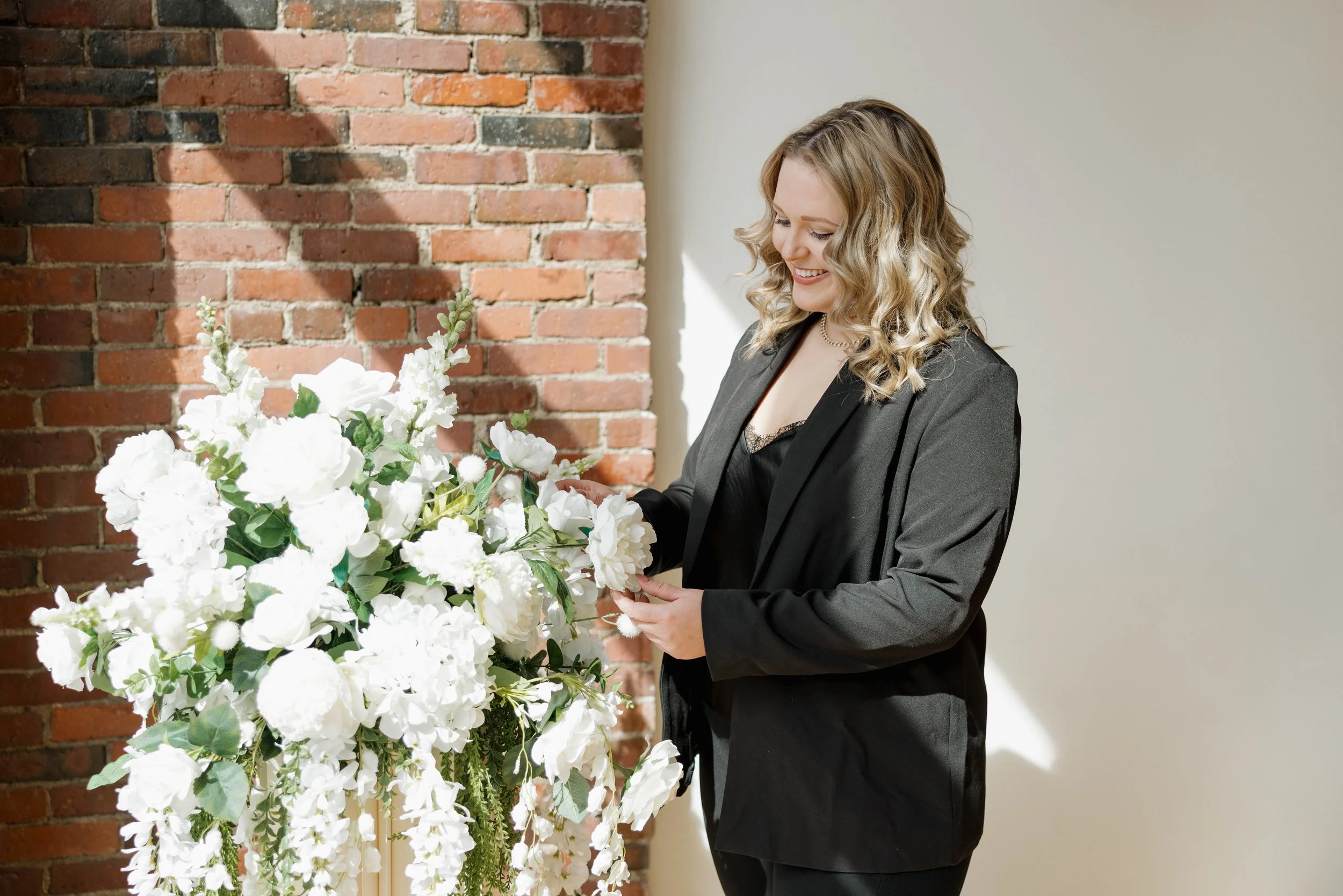A woman with curly blonde hair wearing a black outfit and a gray blazer, smiling as she arranges white flowers in a floral display against a brick wall and a plain wall.