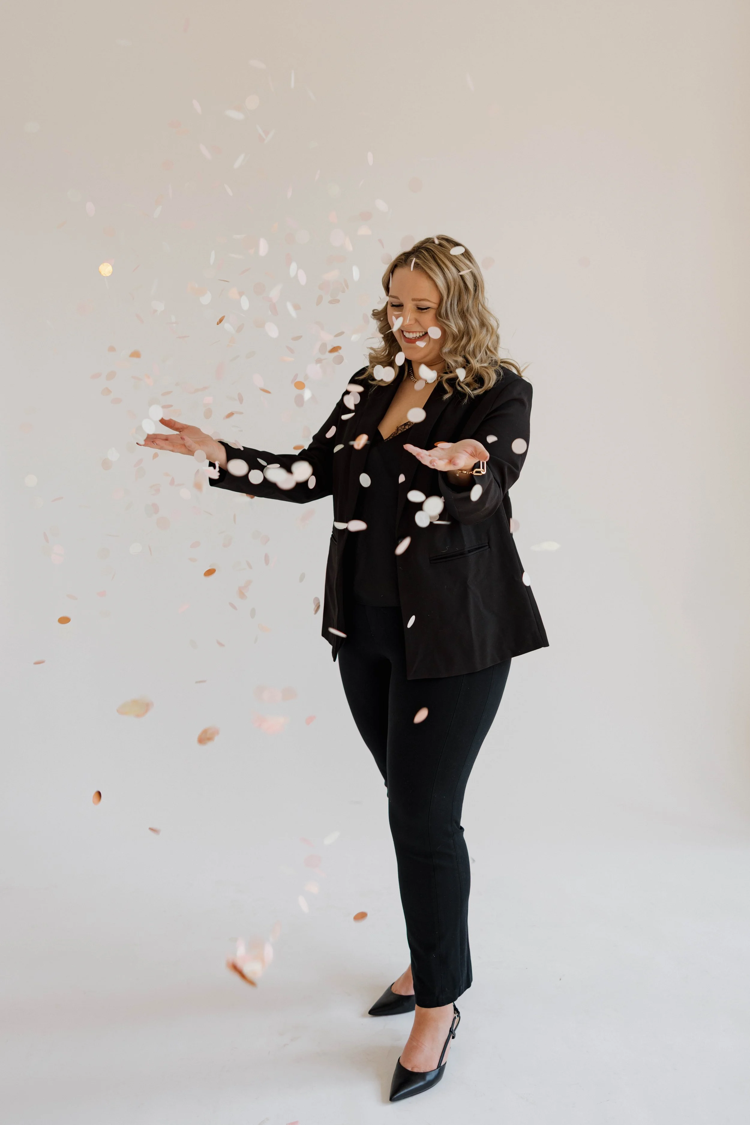 A woman dressed in black pants and a black blazer stands against a plain background while celebrating with falling confetti, smiling joyfully with her eyes closed.