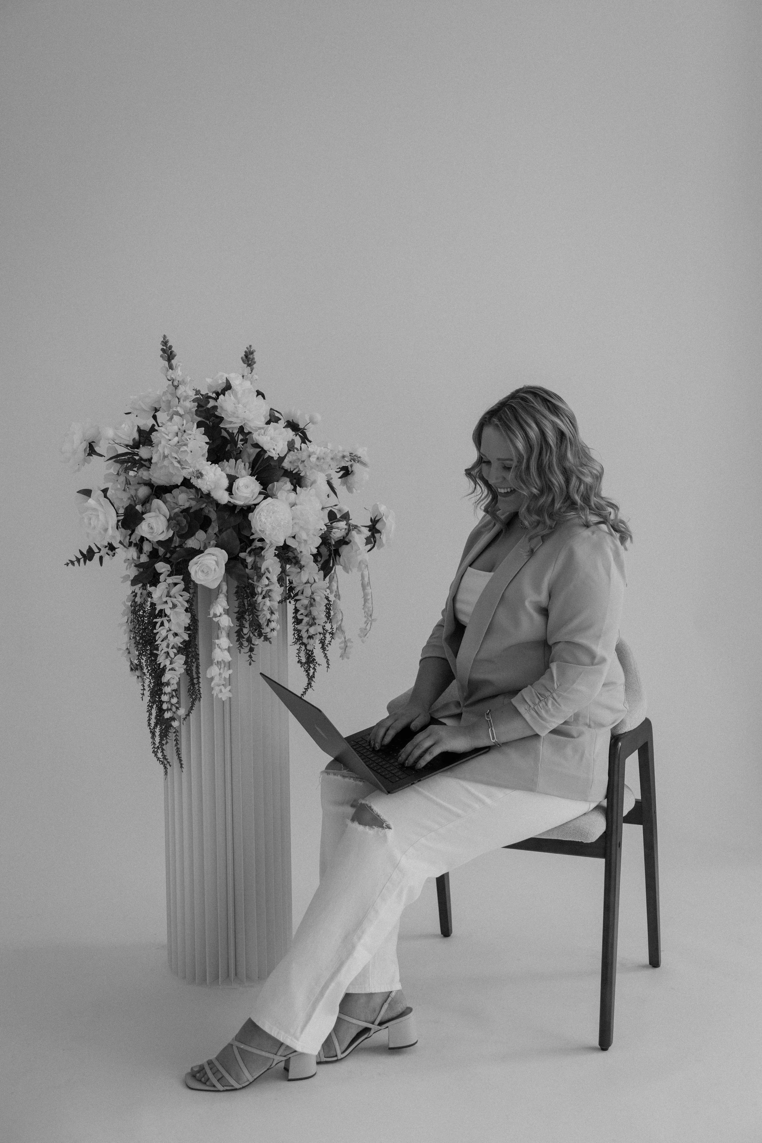 A woman sitting on a chair with a laptop on her lap, smiling. Next to her is a tall vase with a large arrangement of flowers.