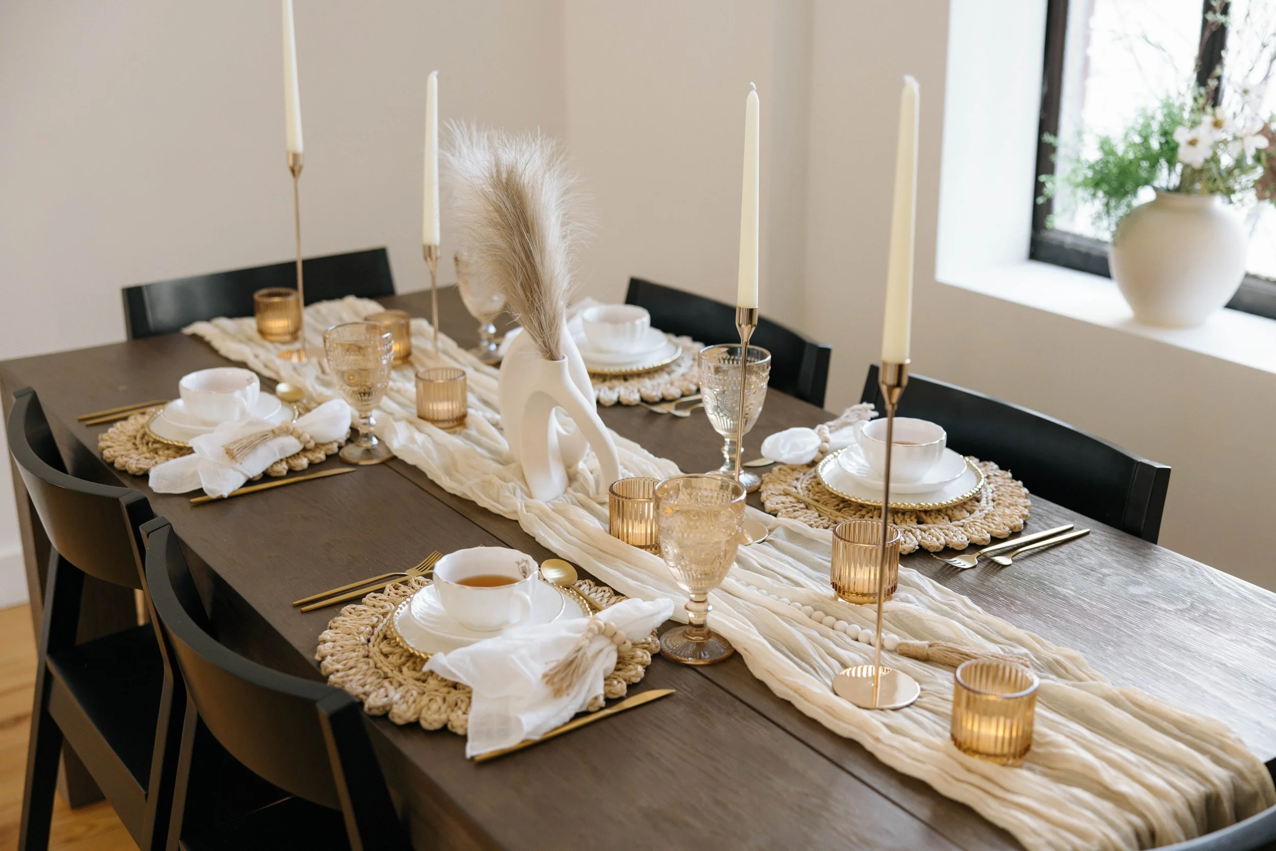 Elegant dining table set for a meal with white plates, tea cups, gold utensils, and a centerpiece with pampas grass. Candles and glassware are also part of the table setting, with a window and a white vase with flowers in the background.