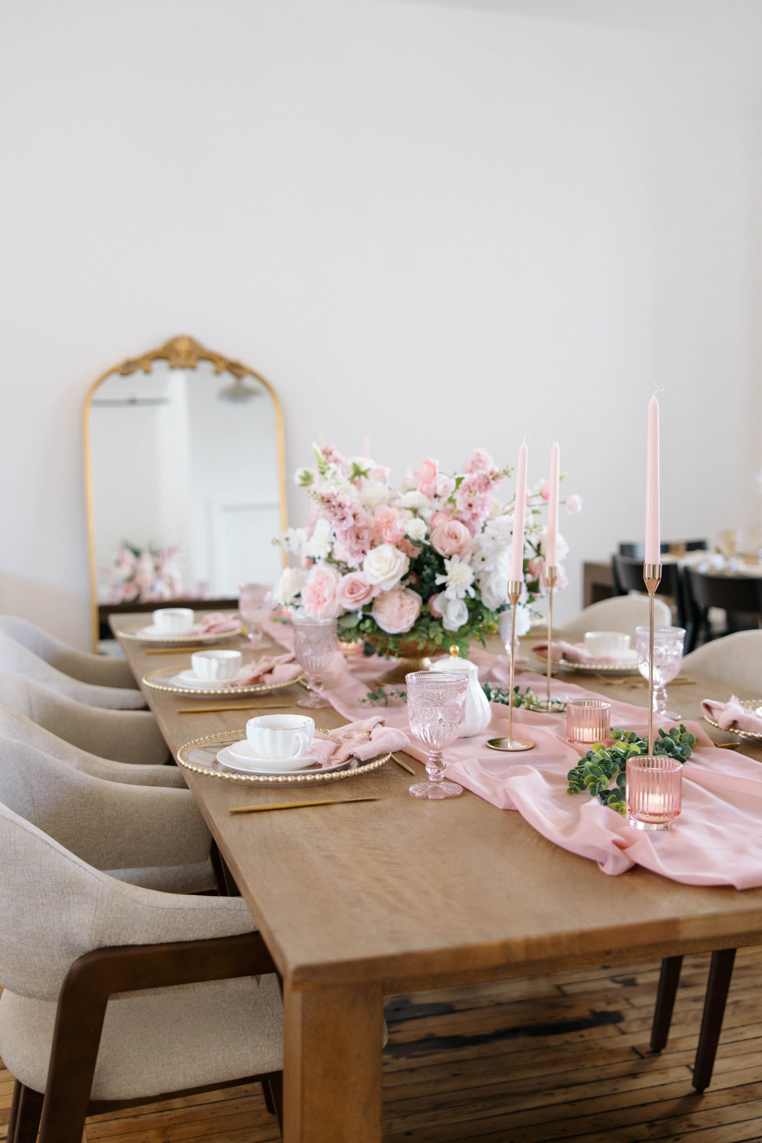 Elegant dining table decorated with pink candles, a large floral centerpiece in shades of pink and white, pink and clear glasses, white plates with pink napkins, and gold utensils, set on a pink runner in a bright room with a large mirror.