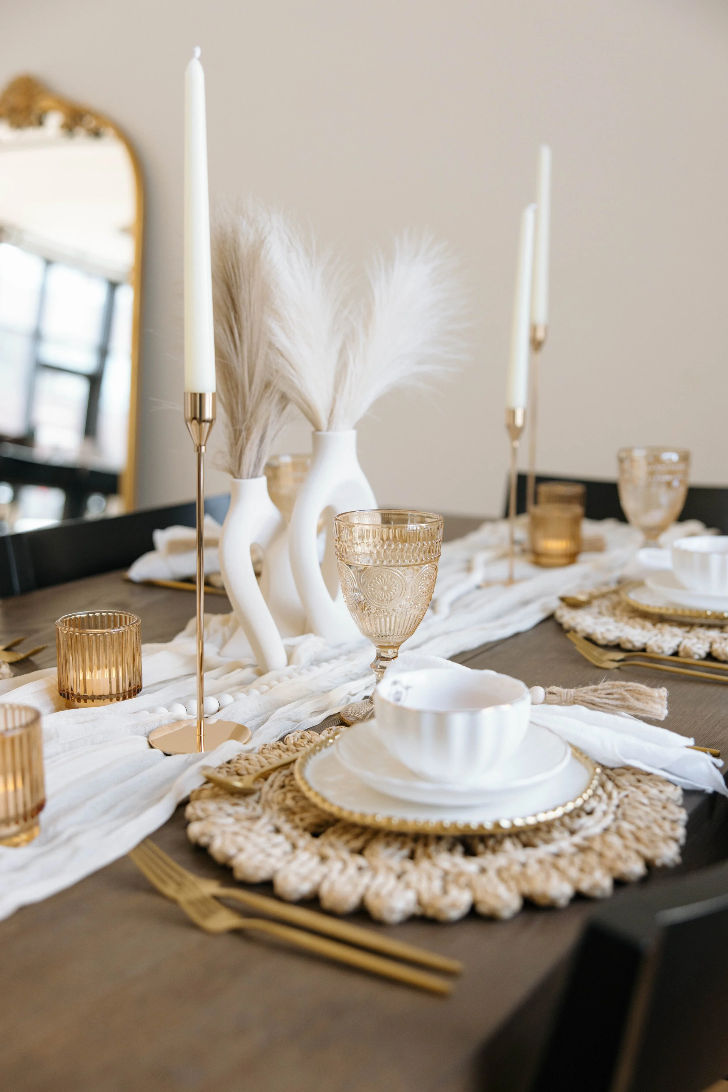 Elegant dining table setting with white and gold accents, including candles, vases with pampas grass, glassware, and napkins.