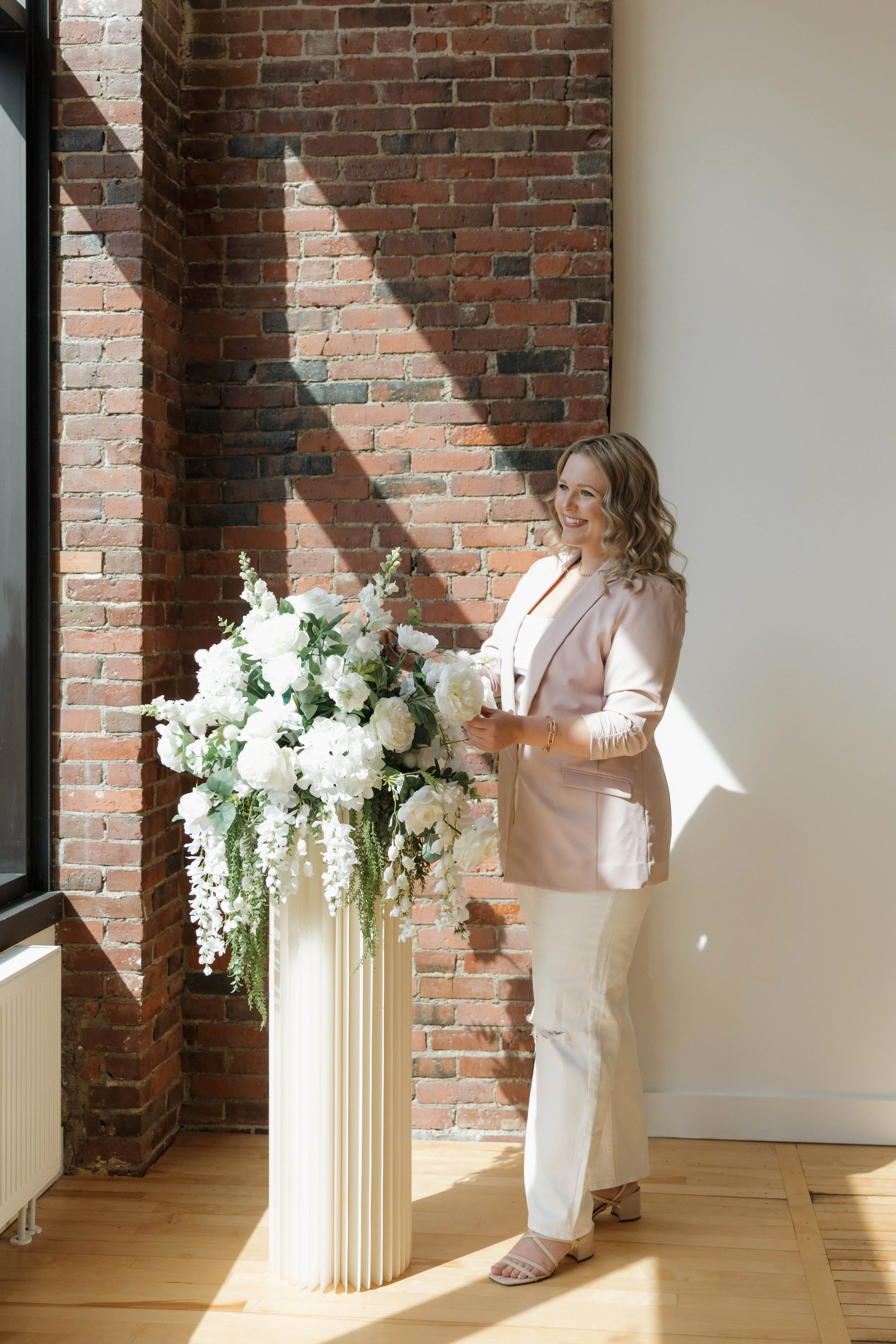 A woman in a light blazer and white pants standing next to a tall cream-colored vase filled with white flowers, in front of a brick wall and a large window letting in sunlight.