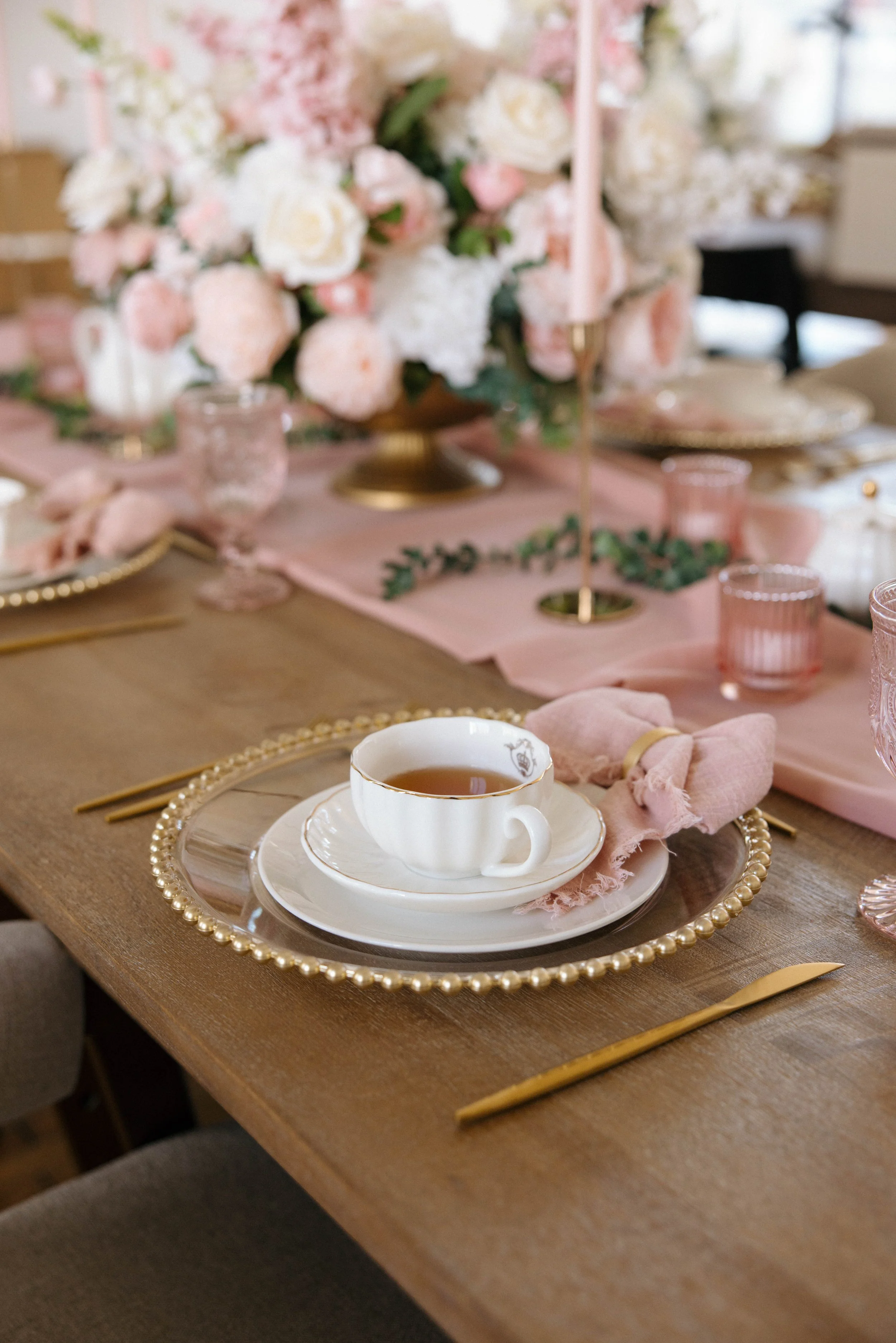 A tea or coffee cup on a plate with a pink napkin, set on a decorative plate with a pearl-like border, at a table with pink and gold accents, floral centerpiece, and pink glasses for a festive event.