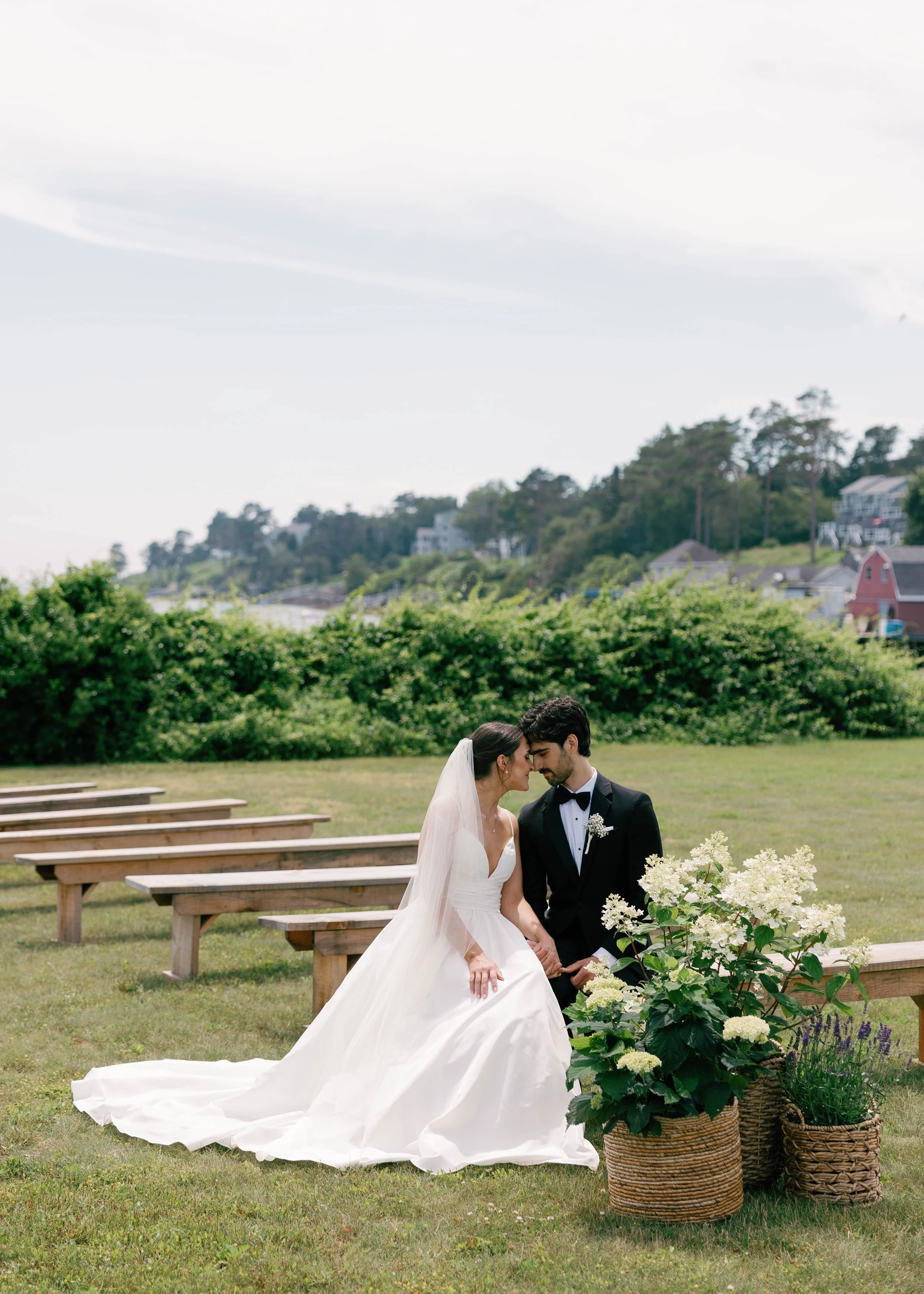 A bride and groom sit together in a grassy outdoor setting during their wedding, surrounded by potted white flowering plants and wooden benches, with green trees and houses on a hillside in the background.