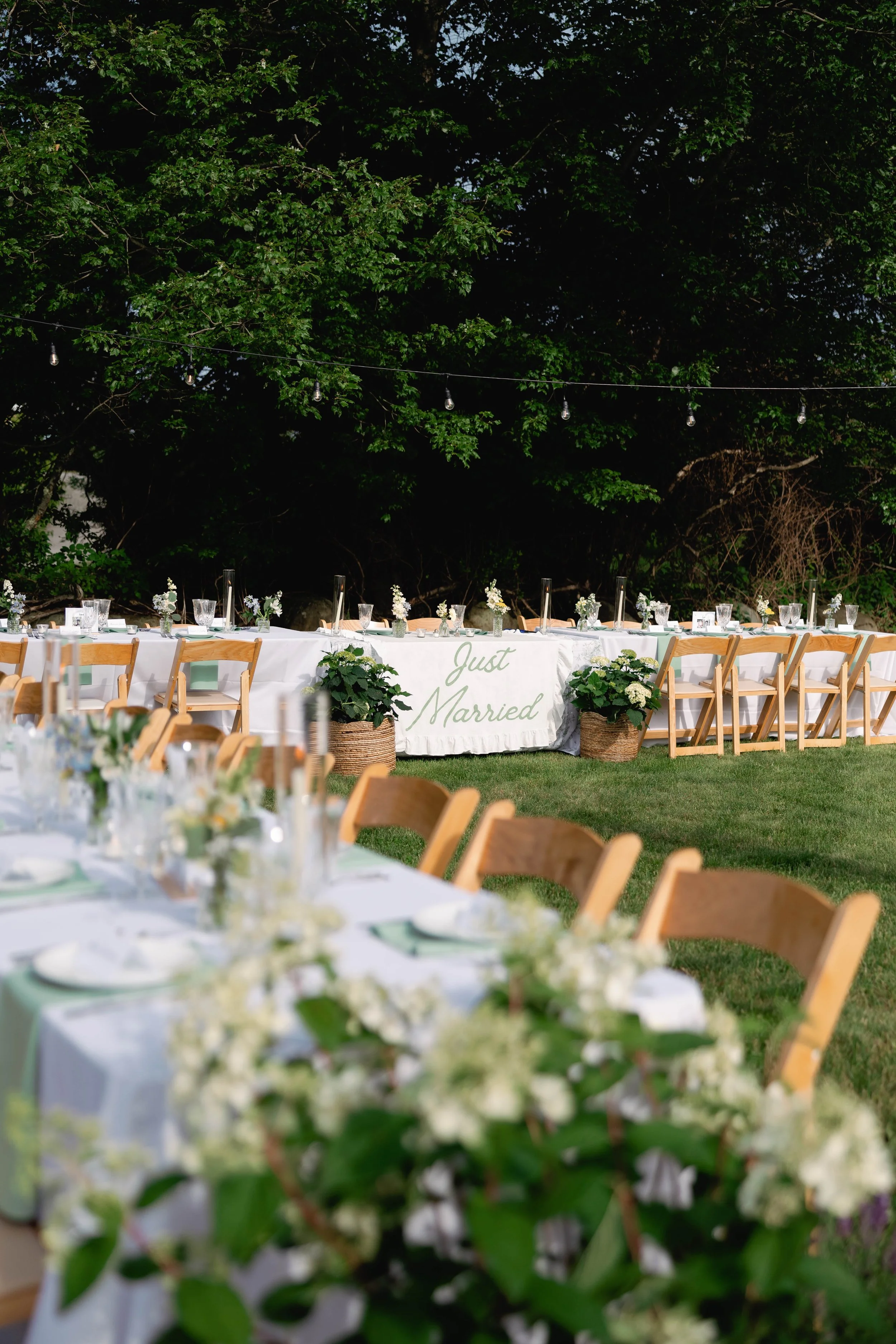 Outdoor wedding reception table decorated with white flowers, greenery, and elegant tableware, with a 'Just Married' sign hanging on the head table.