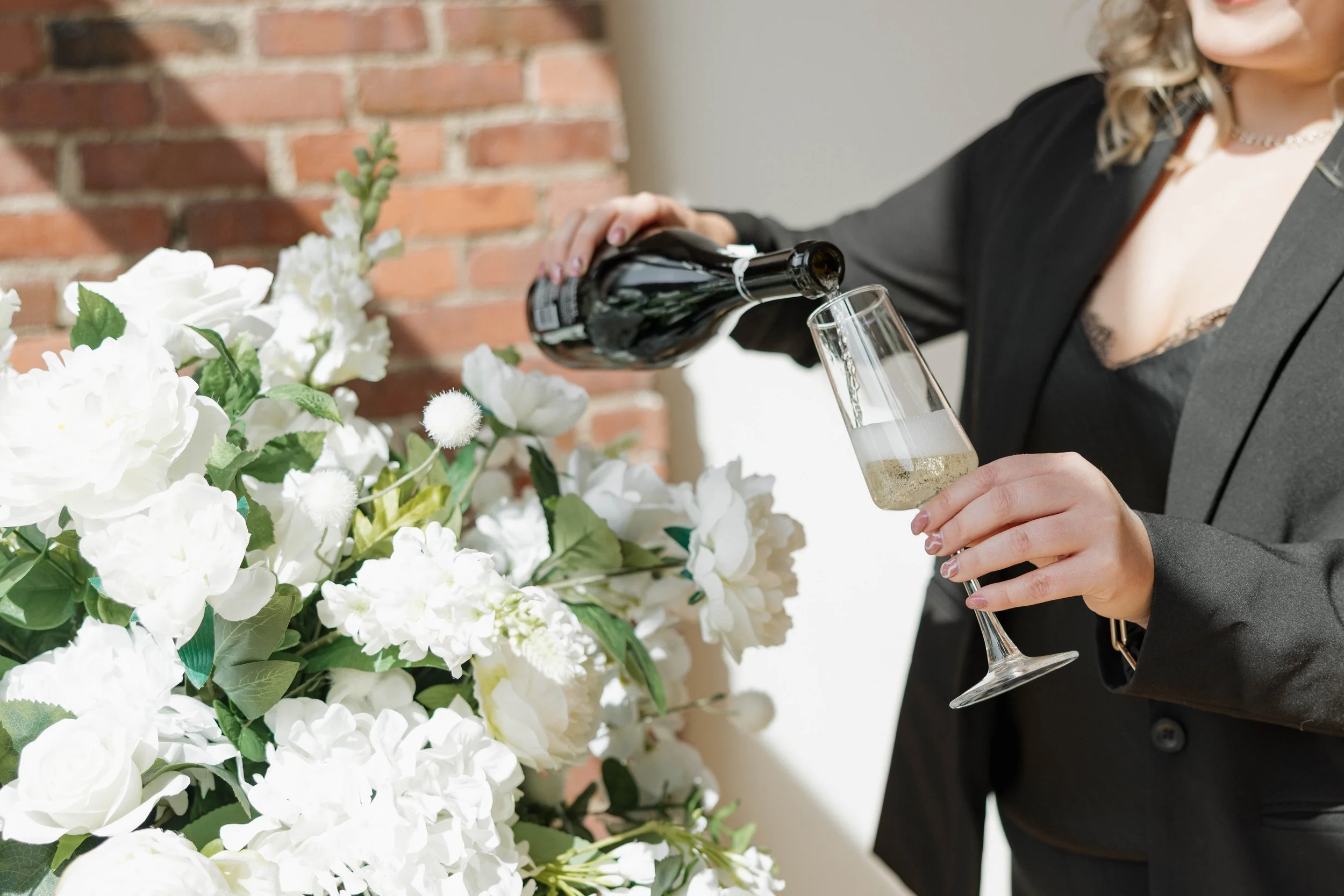 A woman in a black blazer is pouring champagne into a flute next to a large arrangement of white flowers.