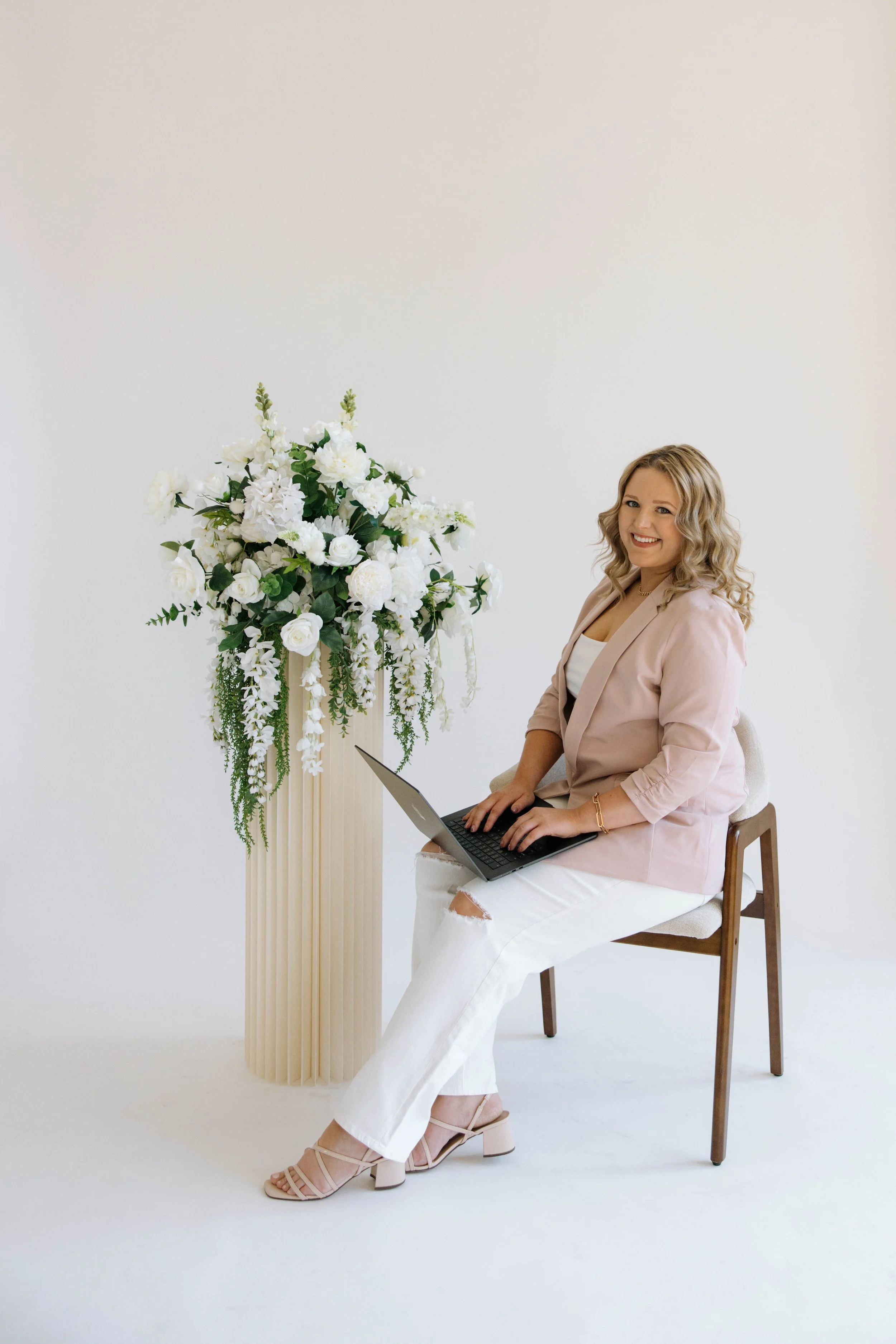 A woman sitting on a chair with a laptop, smiling, near a tall flower arrangement with white flowers and green leaves.