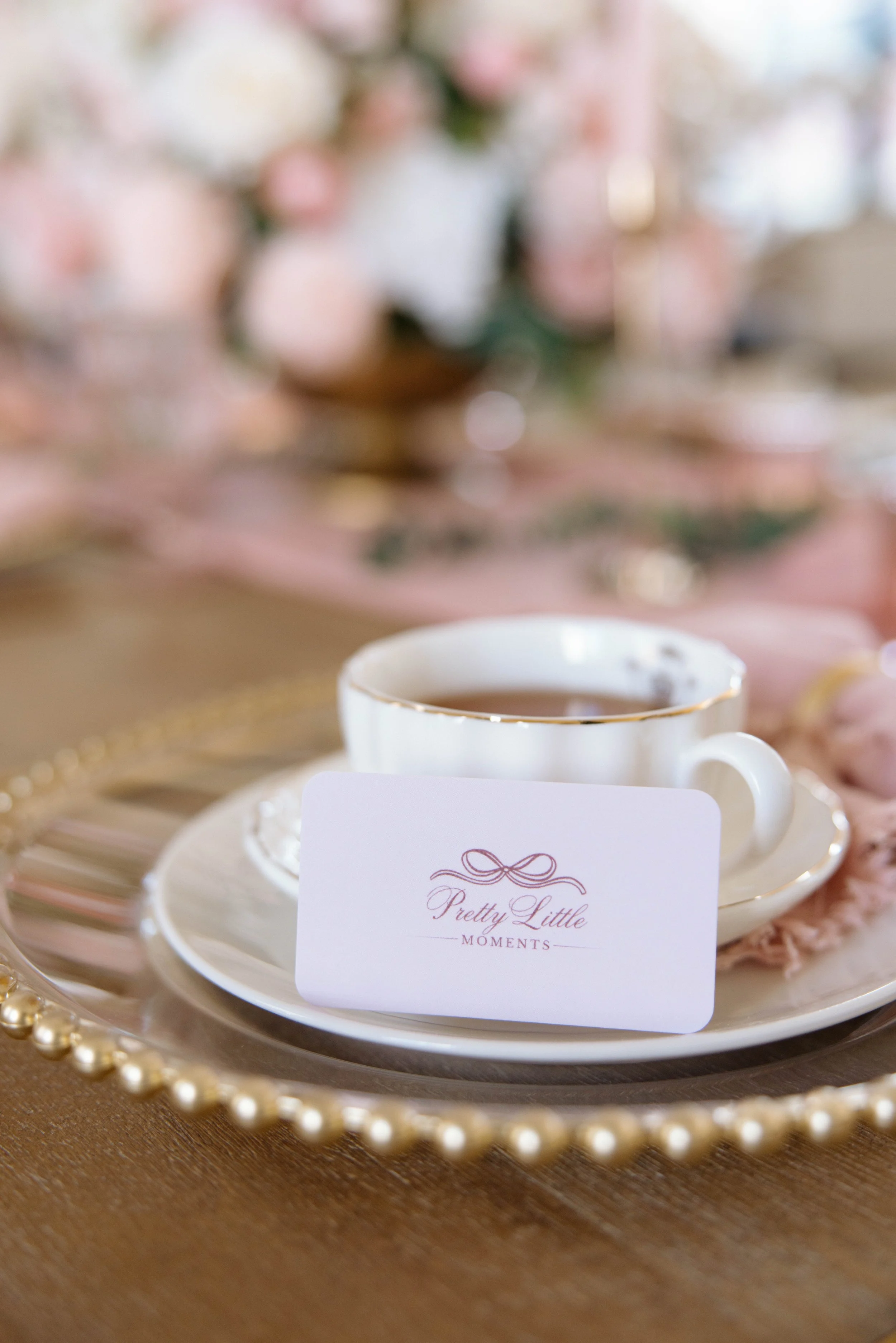 A white teacup filled with tea on a saucer, placed on a decorative golden tray with pearl accents. A place card with elegant script reading 'Pretty Little Moments' is leaning against the teacup. The background is softly blurred with pink and white flowers.
