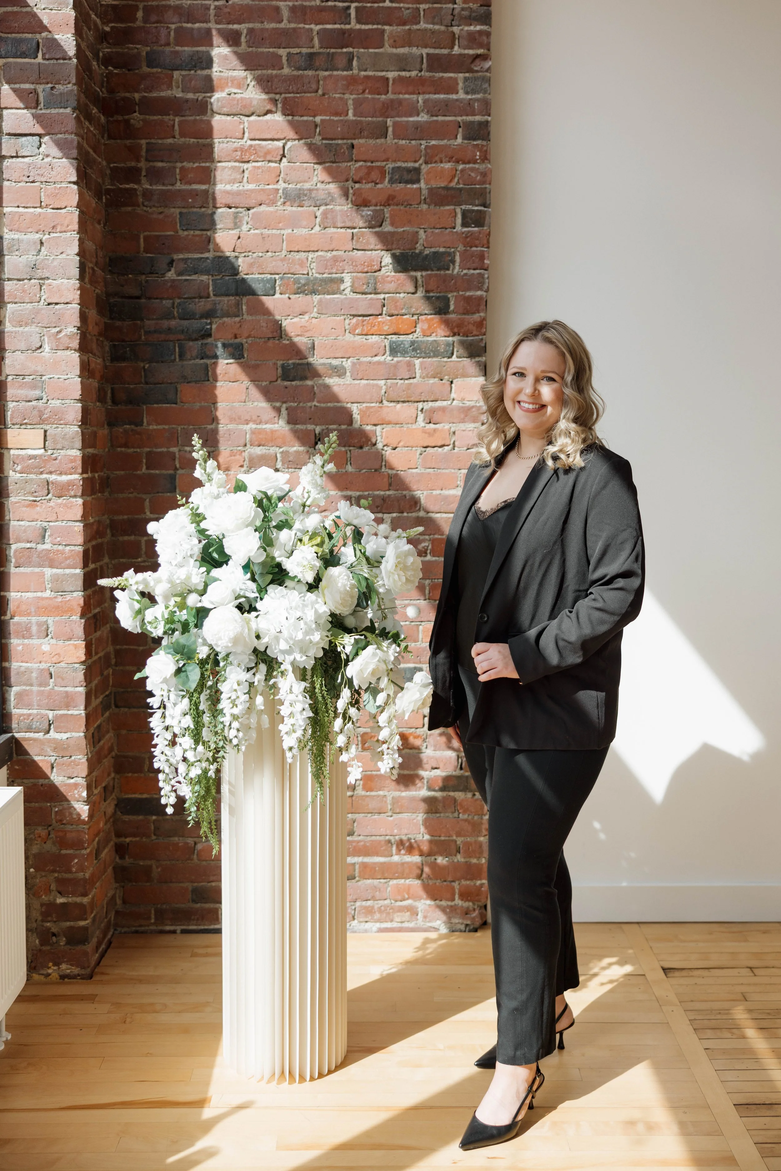 A woman with blonde curly hair dressed in a black blazer, black pants, and black heels standing next to a large white floral arrangement on a tall vase, inside a room with a brick wall and a white wall, and sunlight casting shadows.