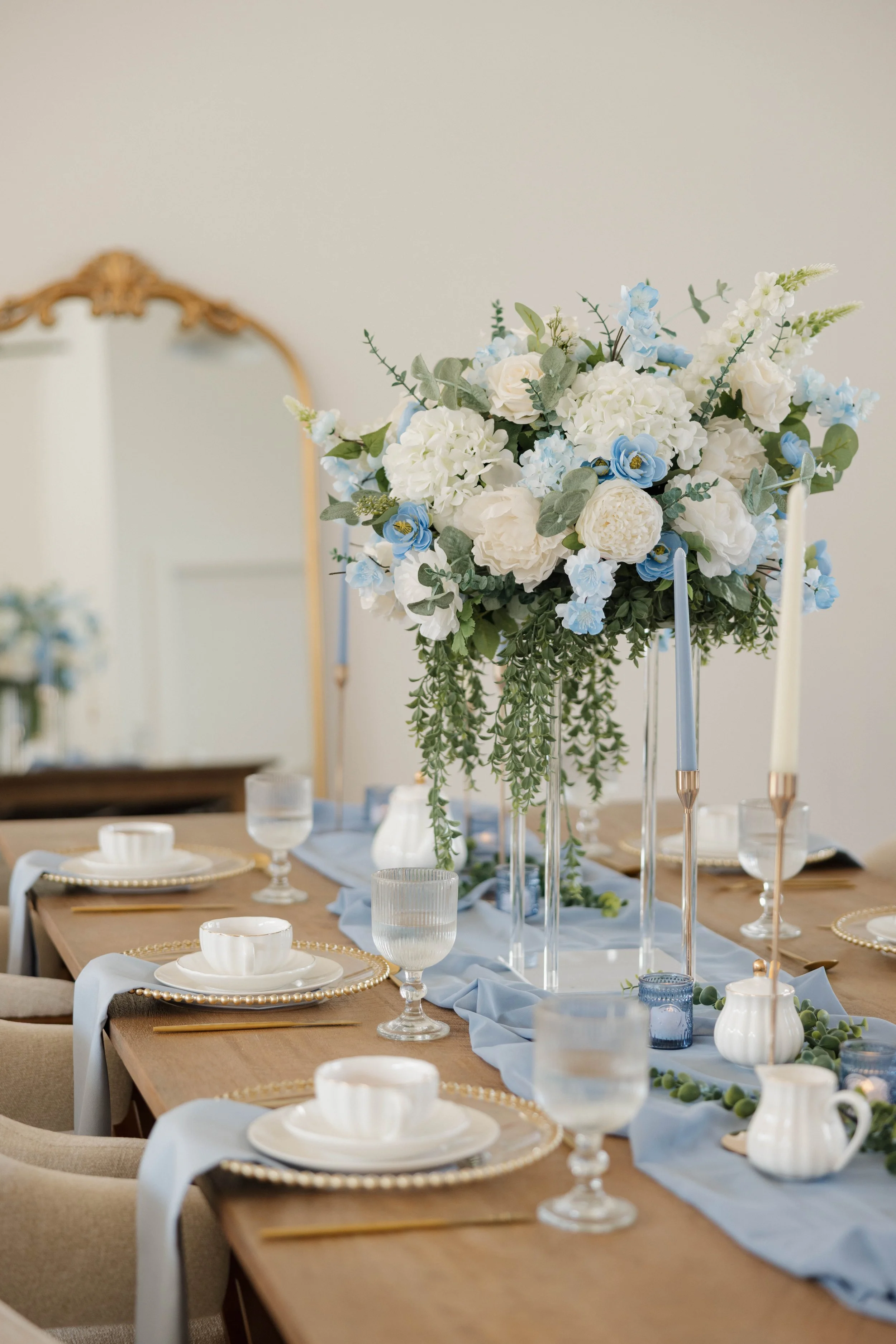 Elegant dining table decorated with a large floral centerpiece of white and light blue flowers, surrounded by white and gold dinnerware, glasses, and tall candlesticks with light-colored candles. A light blue table runner enhances the setting.