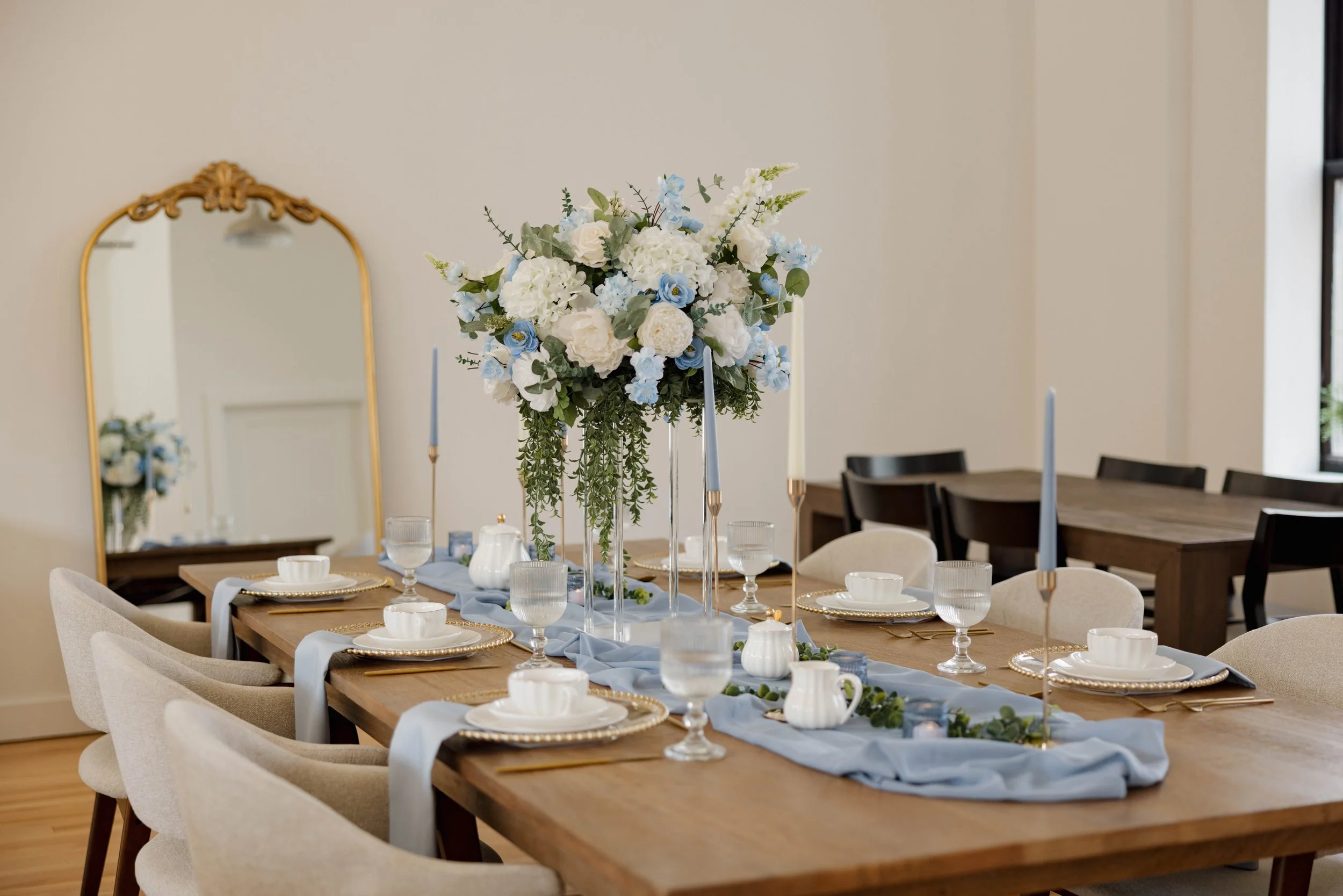 Elegant dining table set with a large floral centerpiece, candles, white china, and glassware, decorated with a blue table runner for a special occasion in a bright room with a mirror on the wall.