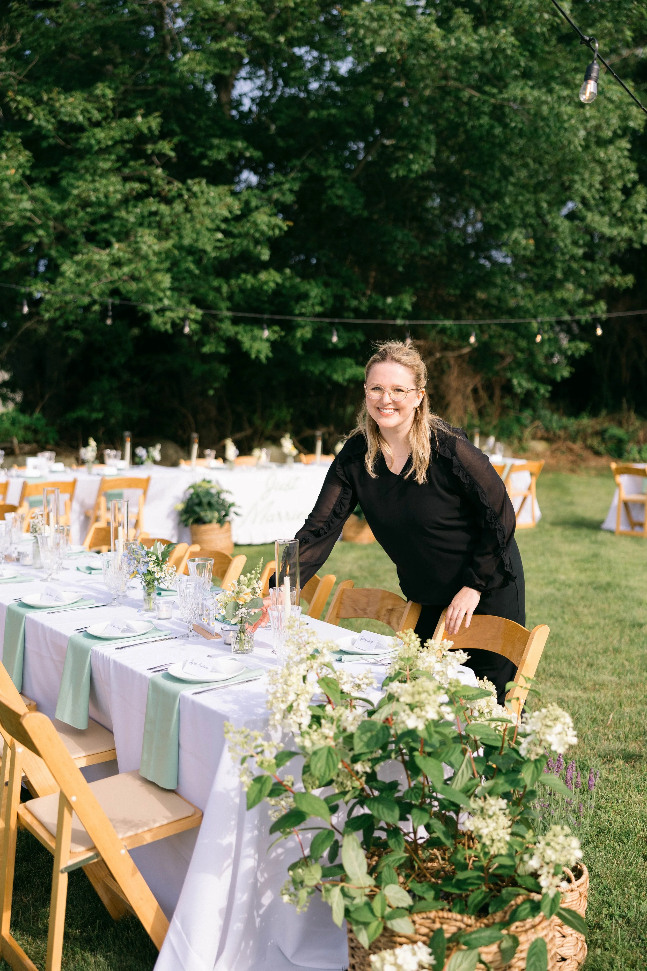 Outdoor wedding reception setup with a long table decorated with floral arrangements, set with plates and glasses, surrounded by wooden chairs. A woman in black attire is smiling and standing at the table, with a lush green trees background and string lights overhead.