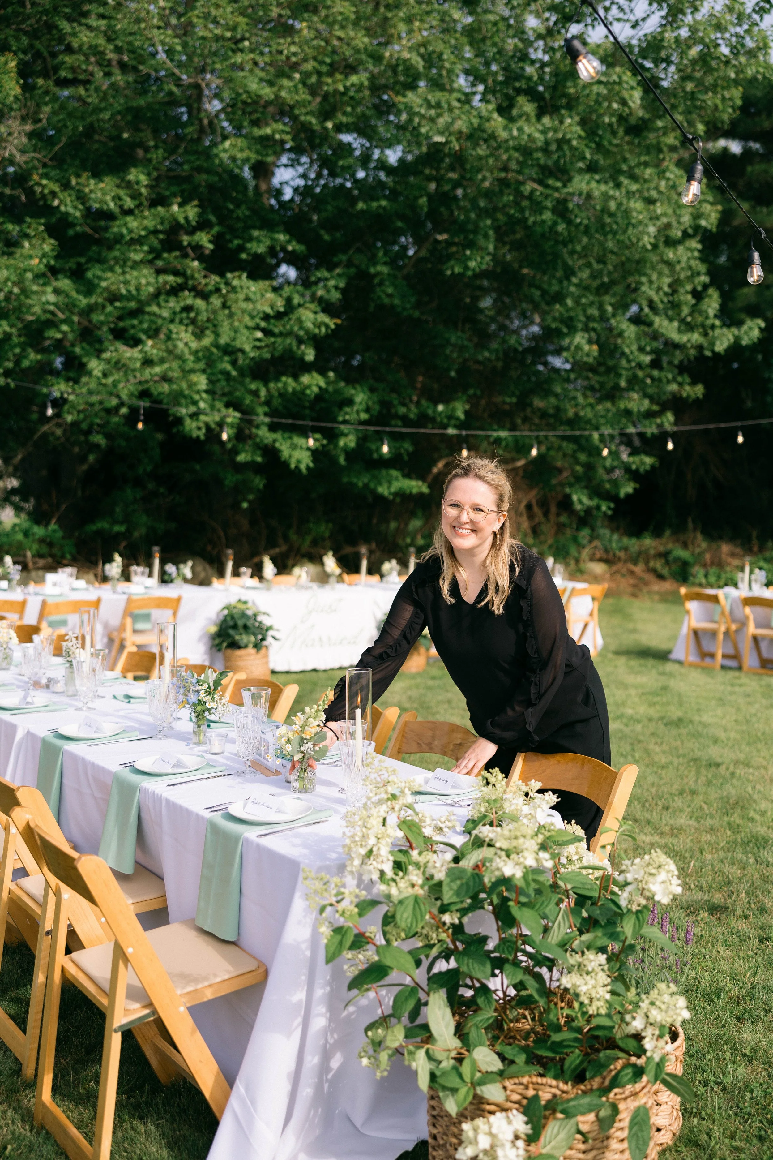 Outdoor reception with a long table decorated with white tablecloths, floral centerpieces, and place cards, set in a garden area with lush green trees and string lights overhead. A woman in black attire is arranging the table, smiling at the camera.