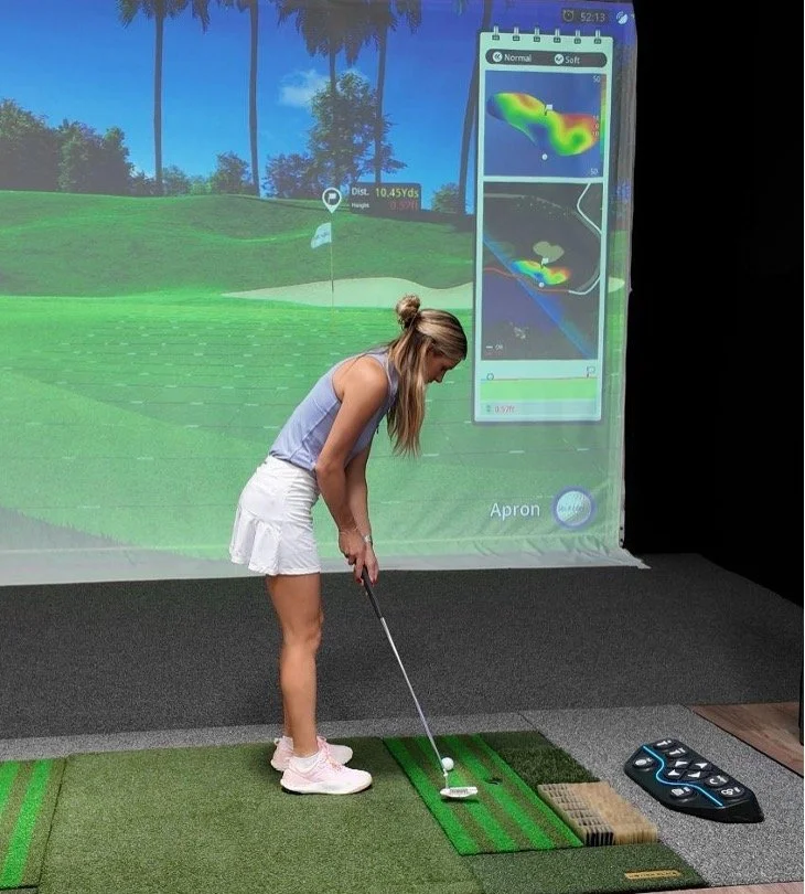 Woman at indoor golf simulator, preparing to putt on green mat, with projected golf course and swing analysis on screen behind her.