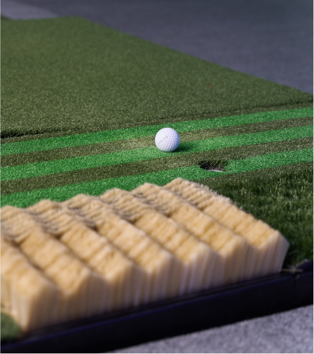Close-up of a golf tee box with a golf ball near the hole on an artificial turf putting green, and a yellow tee on the side.