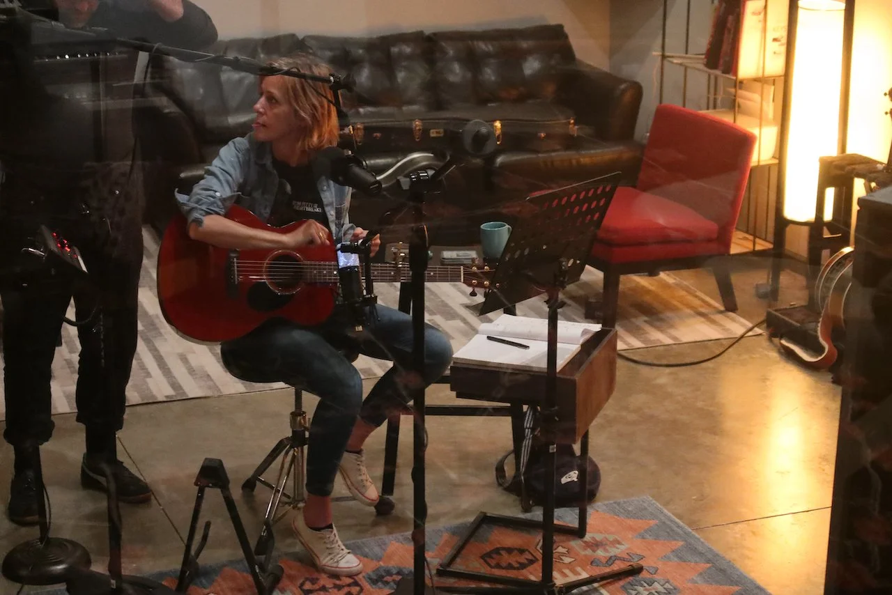 A woman is sitting and playing an acoustic guitar in a cozy room with a brown leather sofa and a red chair. She is surrounded by music equipment, including a microphone, music stand, and a small wooden table with a notebook and pen. There are warm lights and decorative elements like a bookshelf and a patterned rug.
