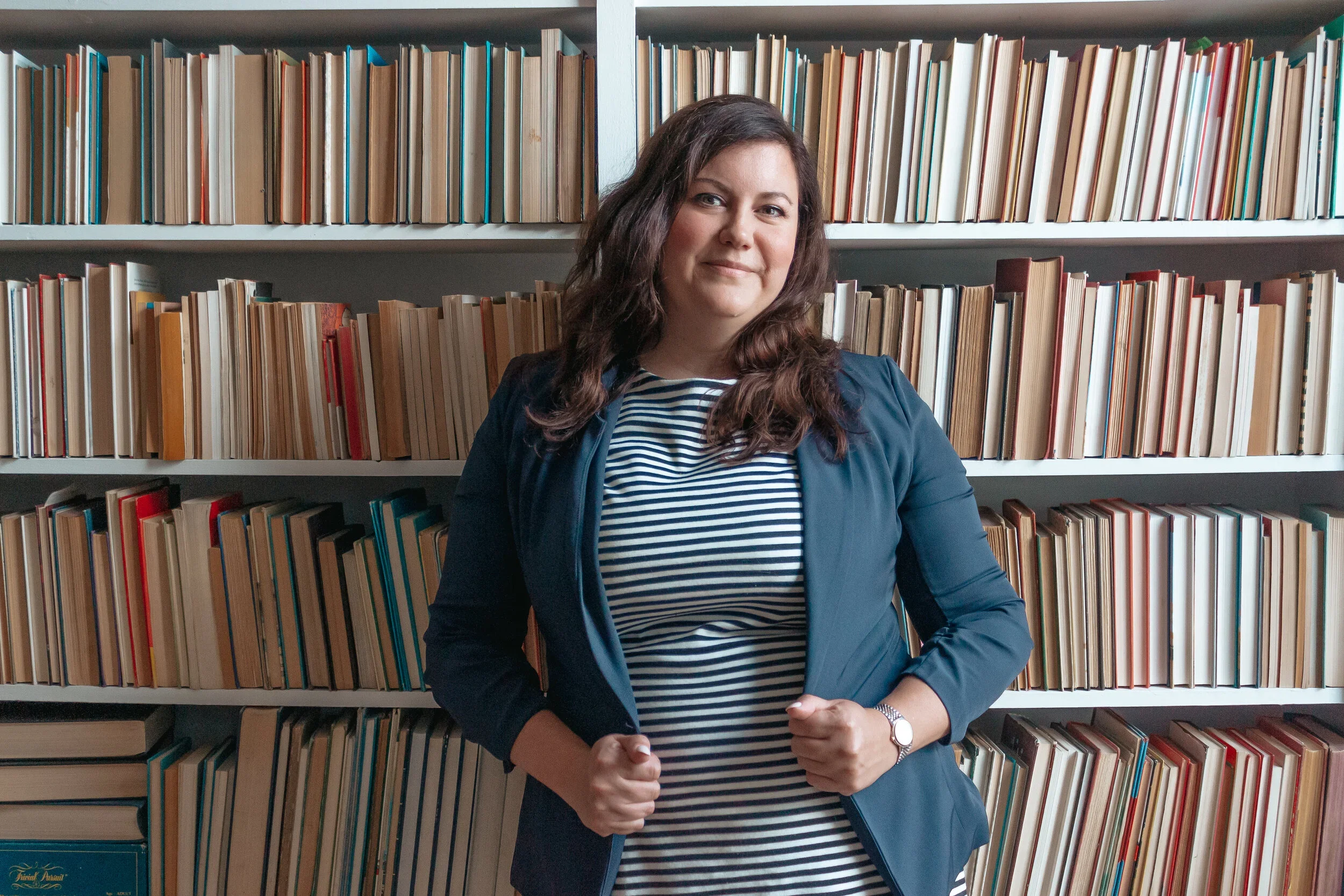 A woman standing in front of bookshelves filled with books, wearing a blue jacket over a striped shirt, with a watch on her left wrist, looking confidently at the camera.