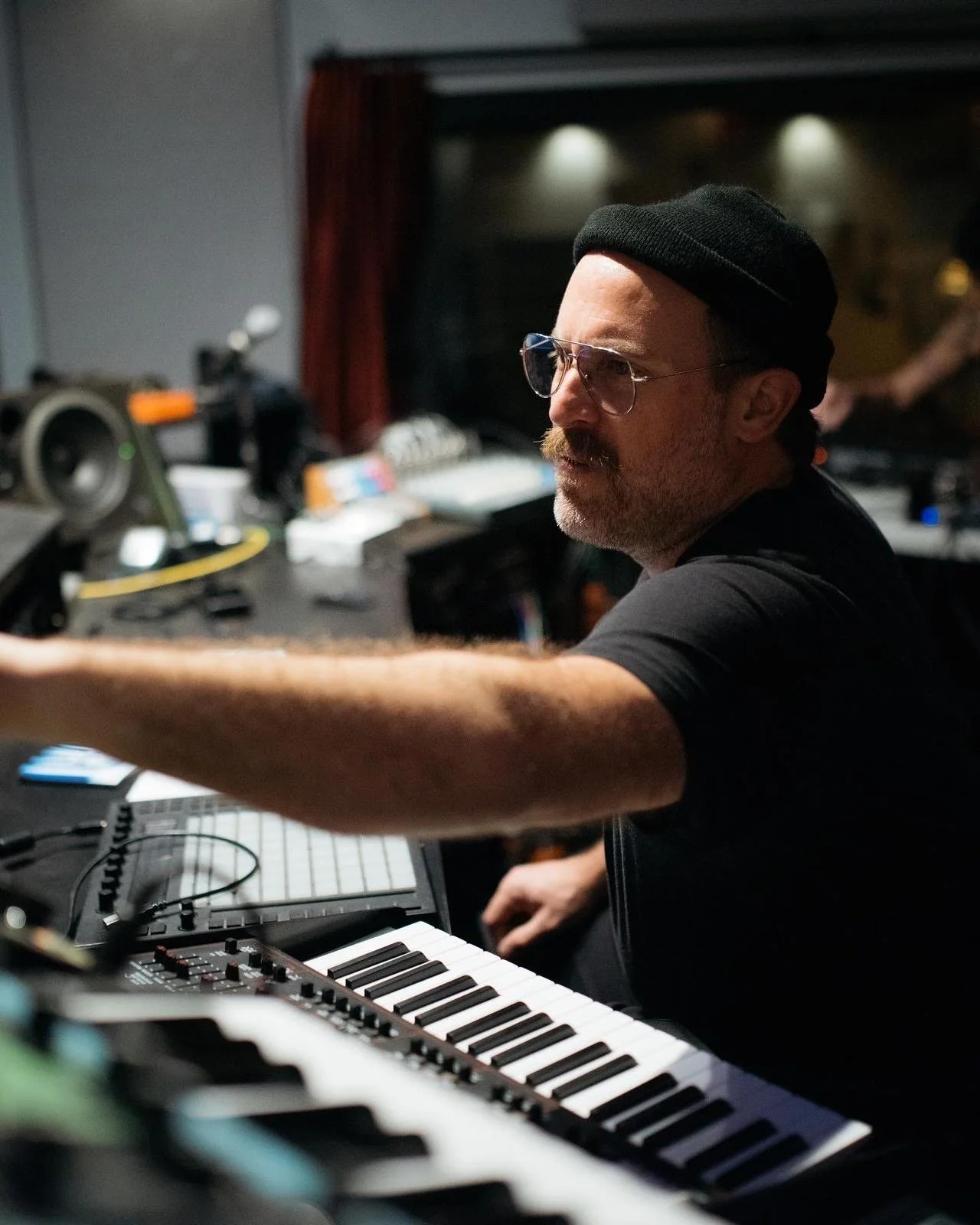 A man with glasses, a black hat, and a beard is playing keyboard in a recording studio.